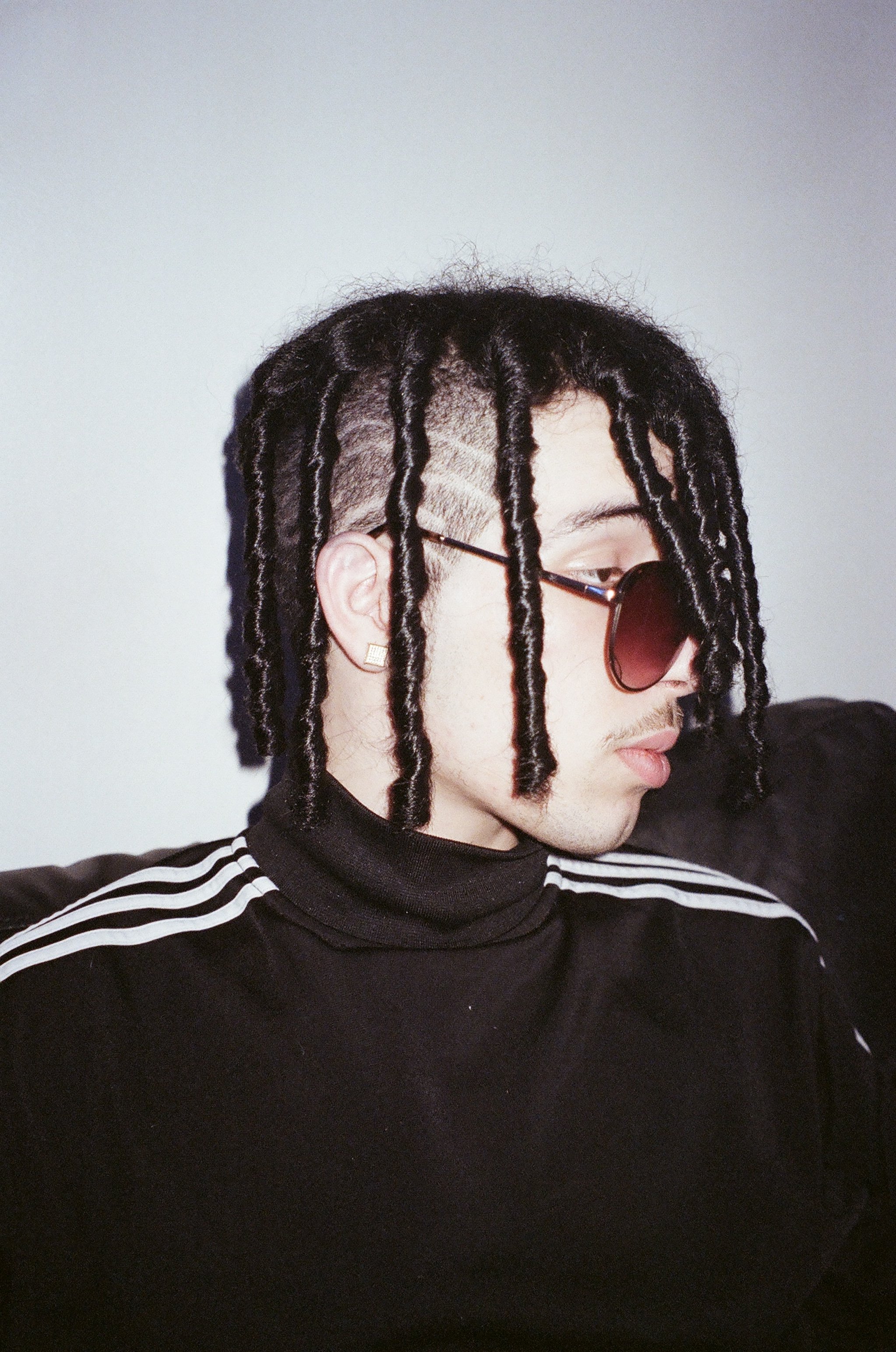 Side profile of a young man with braided dreadlocks, wearing sunglasses and a black turtleneck with white stripes on the shoulders, sitting against a plain wall.