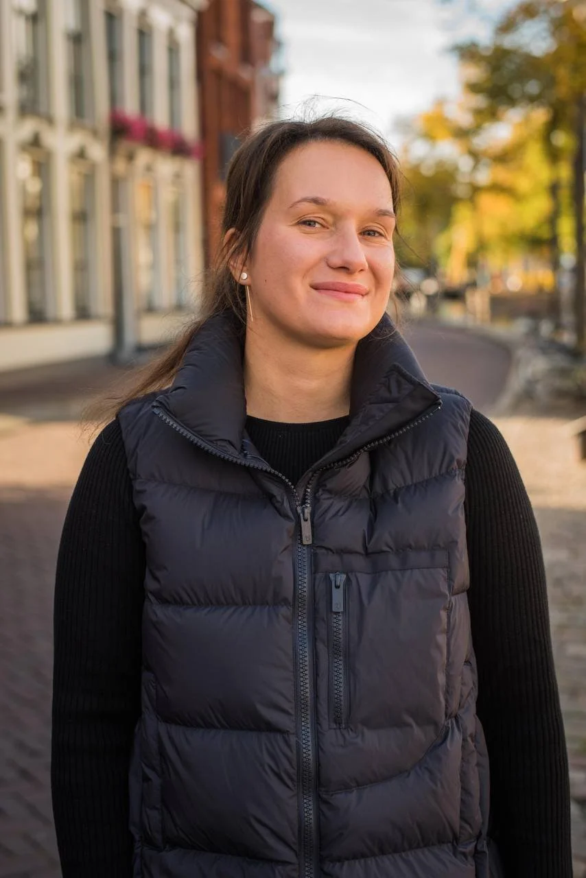 A woman standing outdoors on a cobblestone street, wearing a black puffer vest over a black long-sleeve shirt, with autumn trees and buildings in the background.