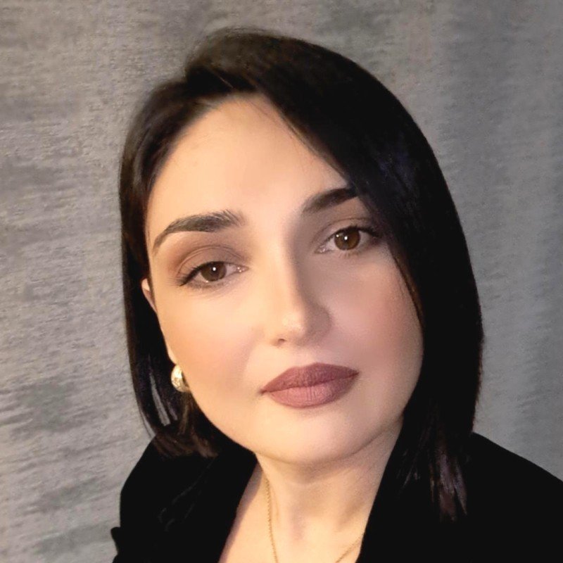 A woman with short dark hair, wearing makeup with lipstick, earrings, and a black top, standing against a textured gray background.