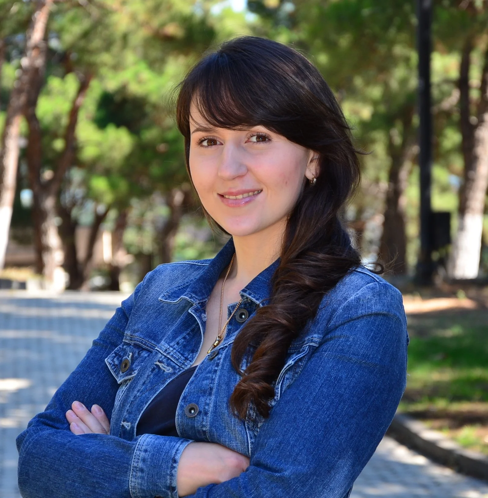 A young woman with long dark hair wearing a denim jacket, standing outdoors in a park with trees in the background.