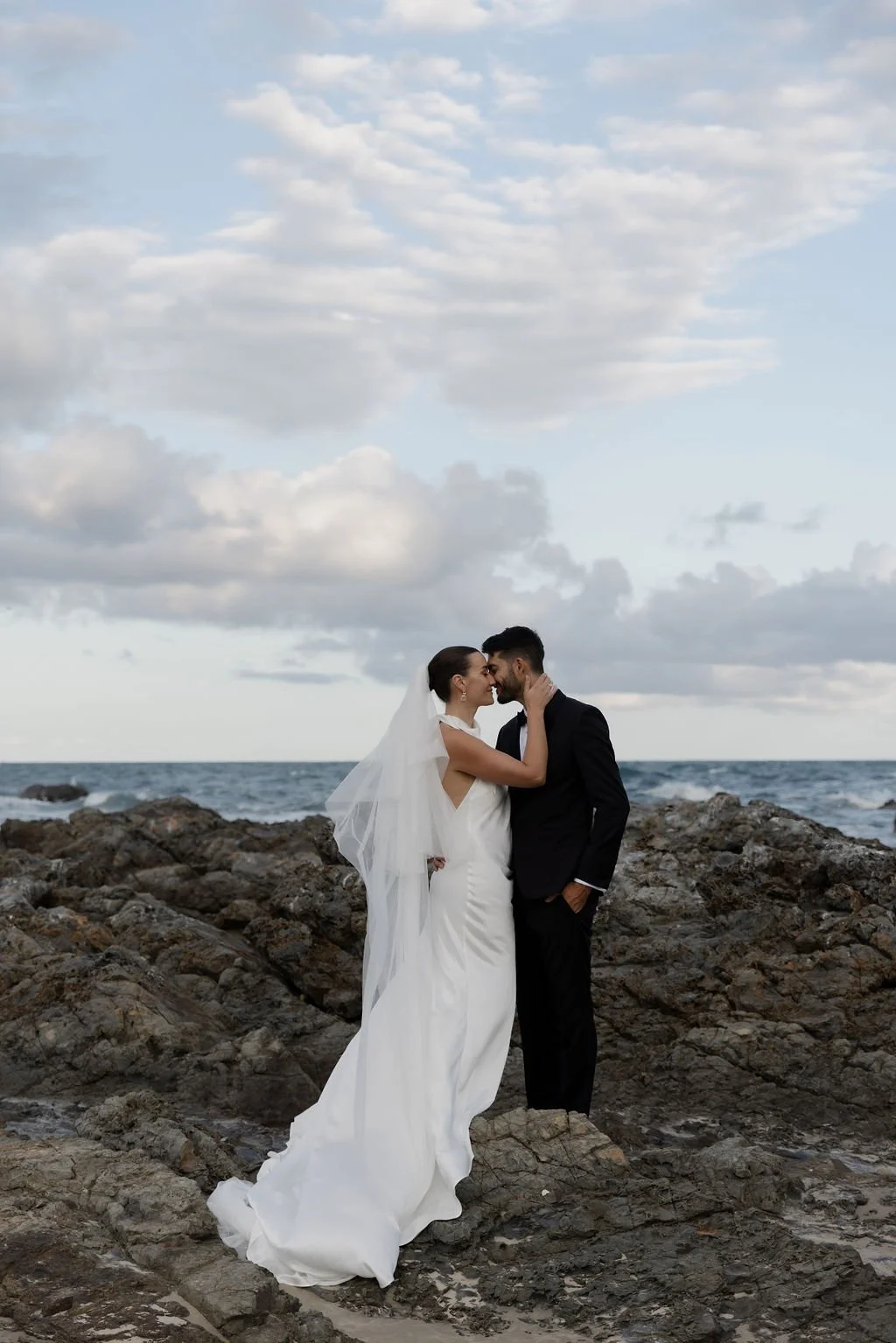 A bride and groom standing on rocks by the ocean, embracing and touching foreheads, under a cloudy sky during sunset.