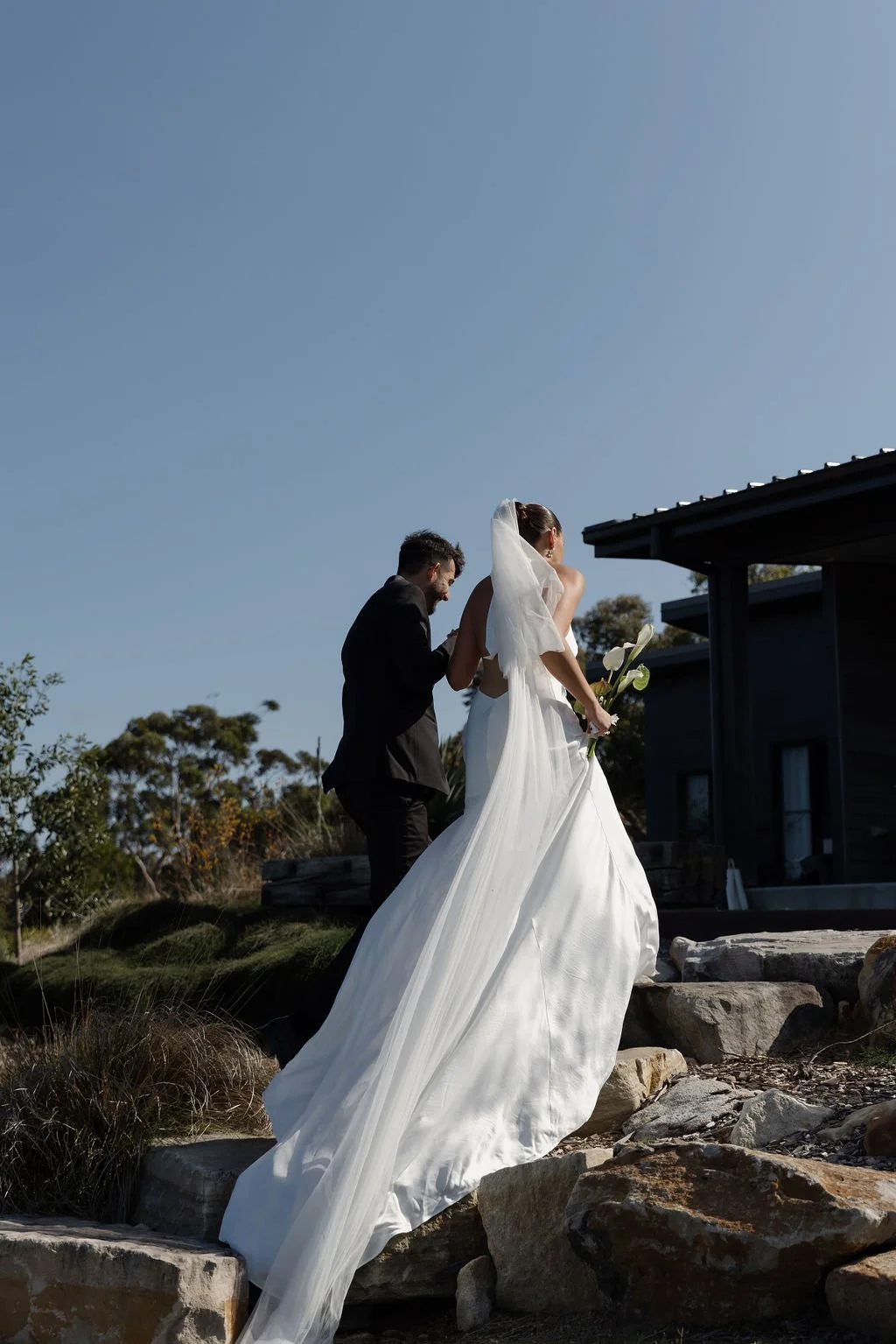 Bride in white wedding gown with long train and veil holding calla lilies, smiling and walking up rocks near a modern house, groom in black suit beside her, outdoors under clear blue sky.