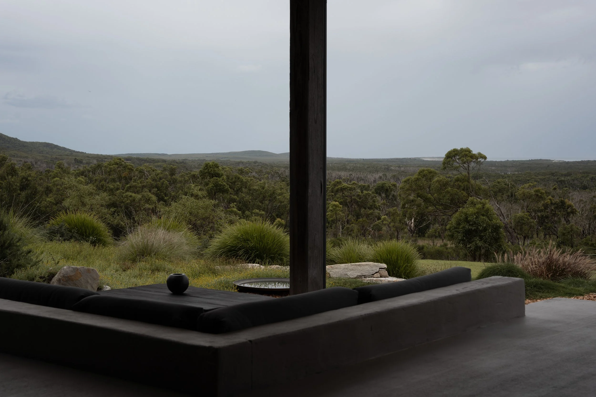 View of a lush forest or nature reserve seen from a shaded porch or covered outdoor seating area, with some decorative objects on the table in the foreground.
