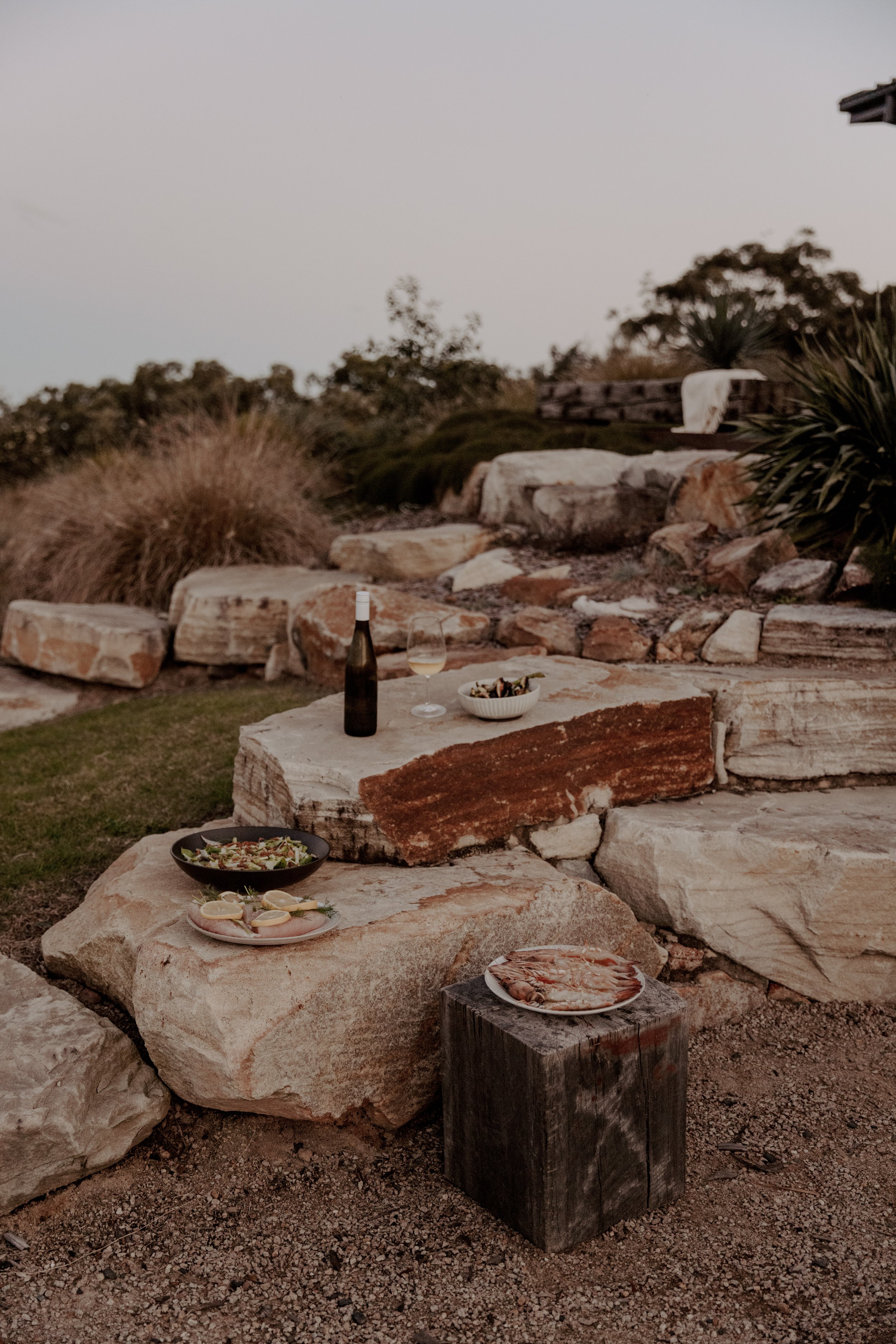 Outdoor dinner setup with a stone table, wine bottle, wine glass, salads, and seafood, with rocks, plants, and trees in the background at sunset.