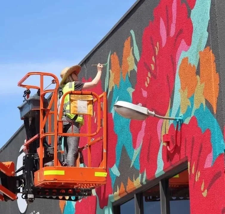 A woman on an elevated orange lift painting a large, colorful mural on the side of a building, under a clear blue sky.