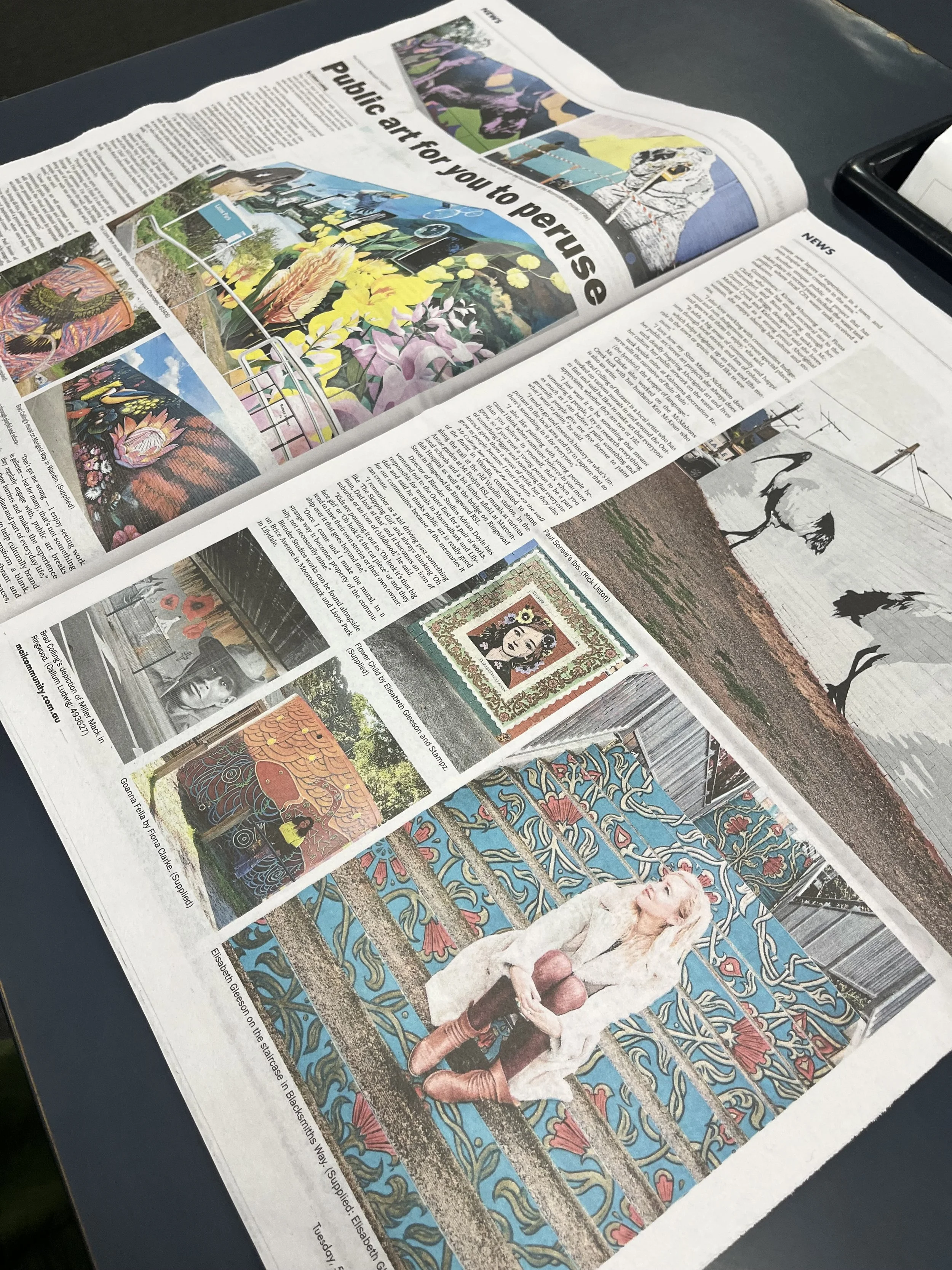 An open newspaper featuring an article about public art for peace, with various colorful images of murals, paintings, and artwork, including a person sitting in front of a vibrant mural.