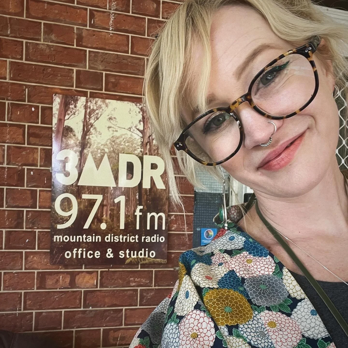 A woman with blonde hair, glasses, and a septum piercing taking a selfie in front of a sign for 3 ADR 97.1 FM Mountain District Radio Office & Studio, which is mounted on a brick wall.