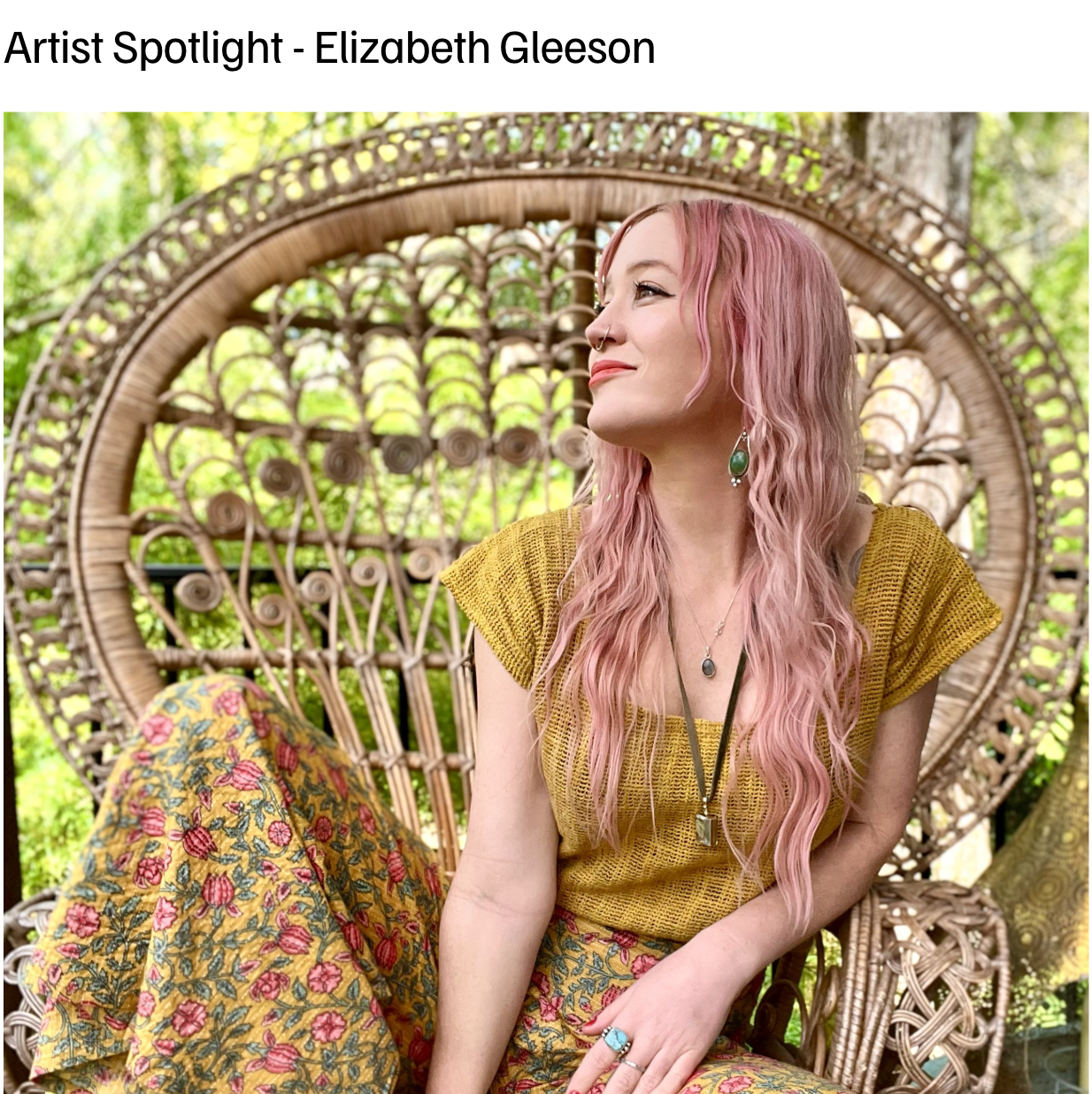 A woman with pink wavy hair, wearing a yellow top and floral pants, sitting on a woven rattan chair outdoors surrounded by greenery.