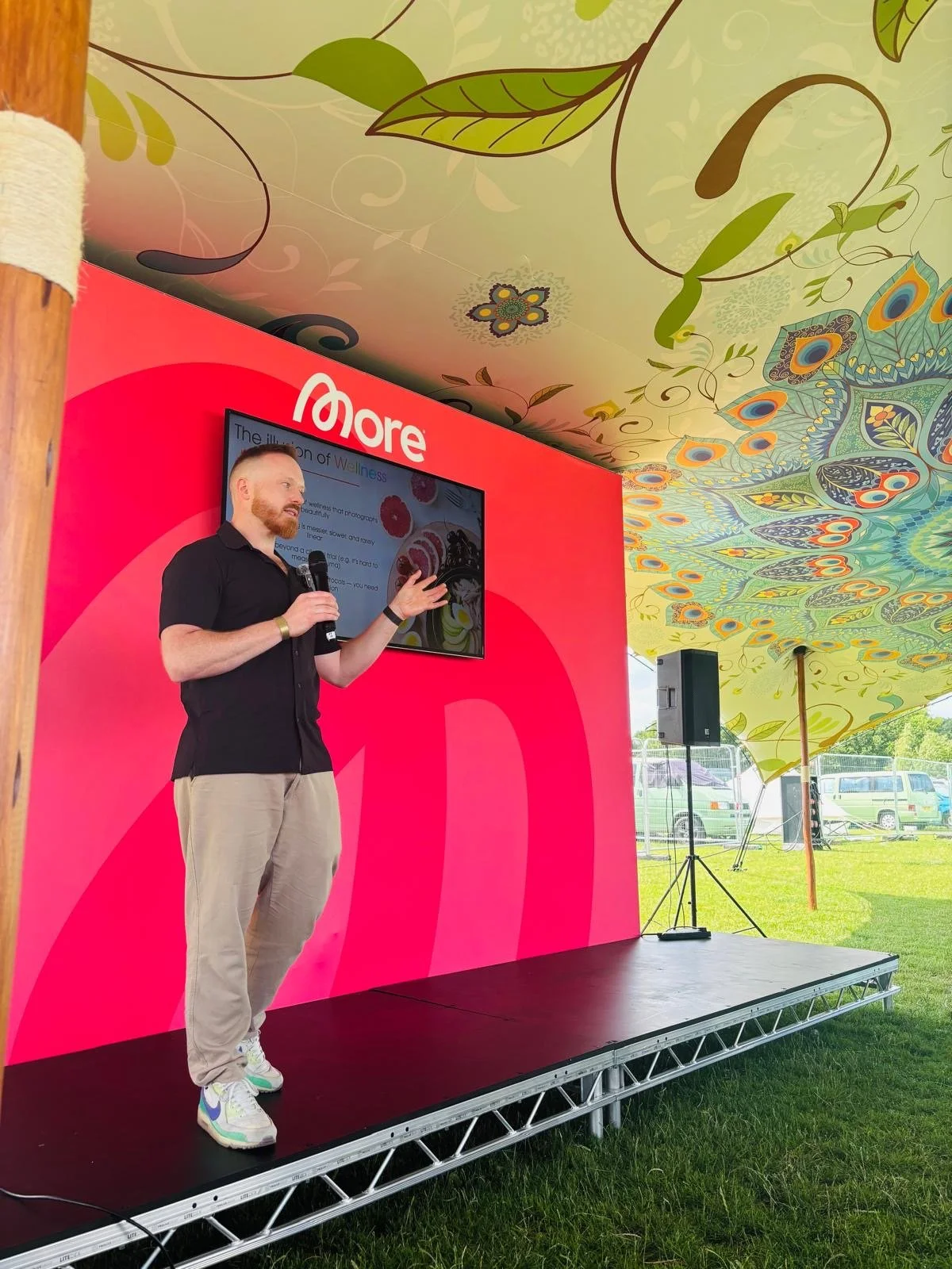 A man with red hair and beard speaking into a microphone on a stage under a colorful, patterned tent at an outdoor event.