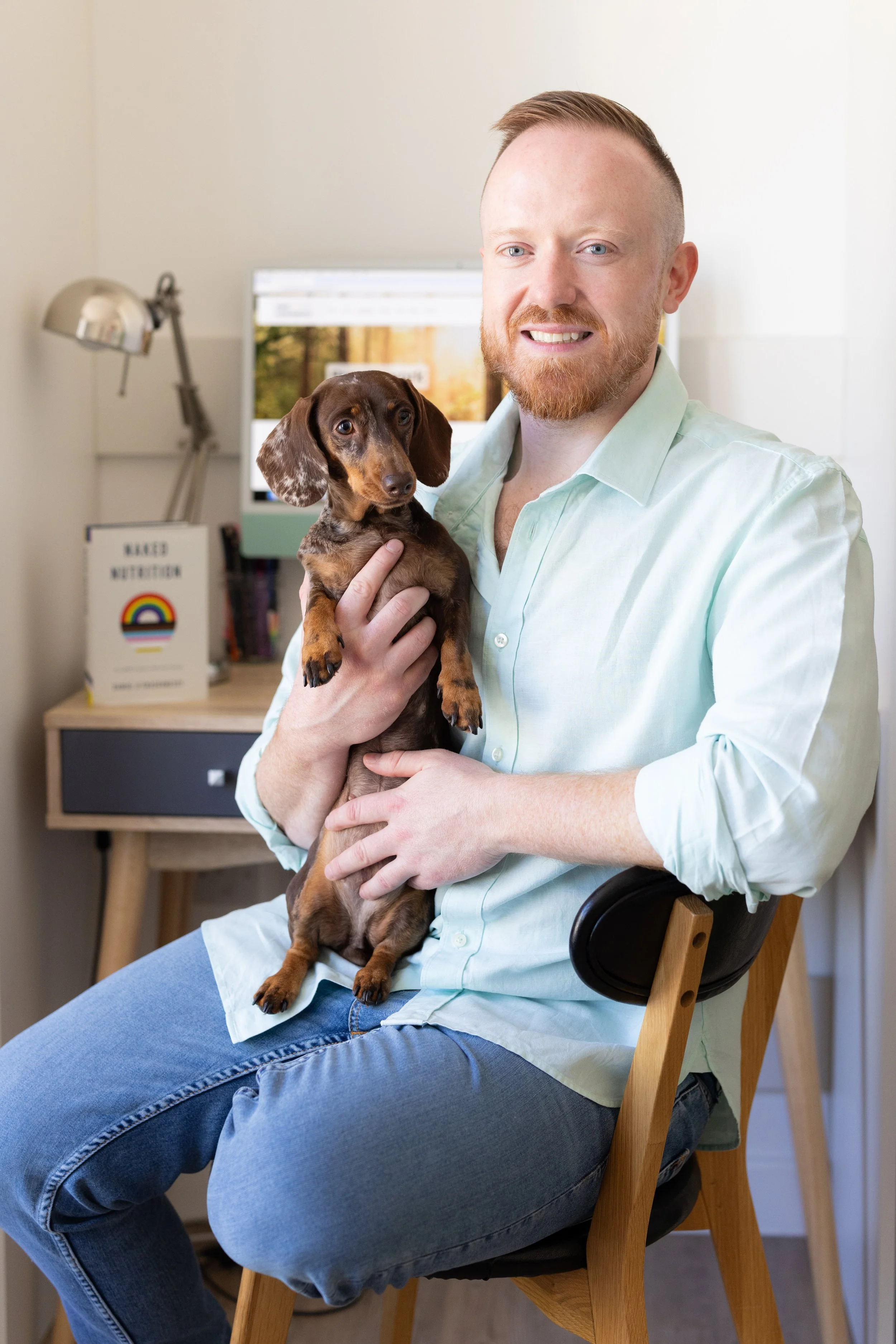 A man with a red beard and light green shirt sitting on a wooden chair, holding a small brown and tan dachshund puppy. In the background, there is a desk with a lamp and a computer monitor.