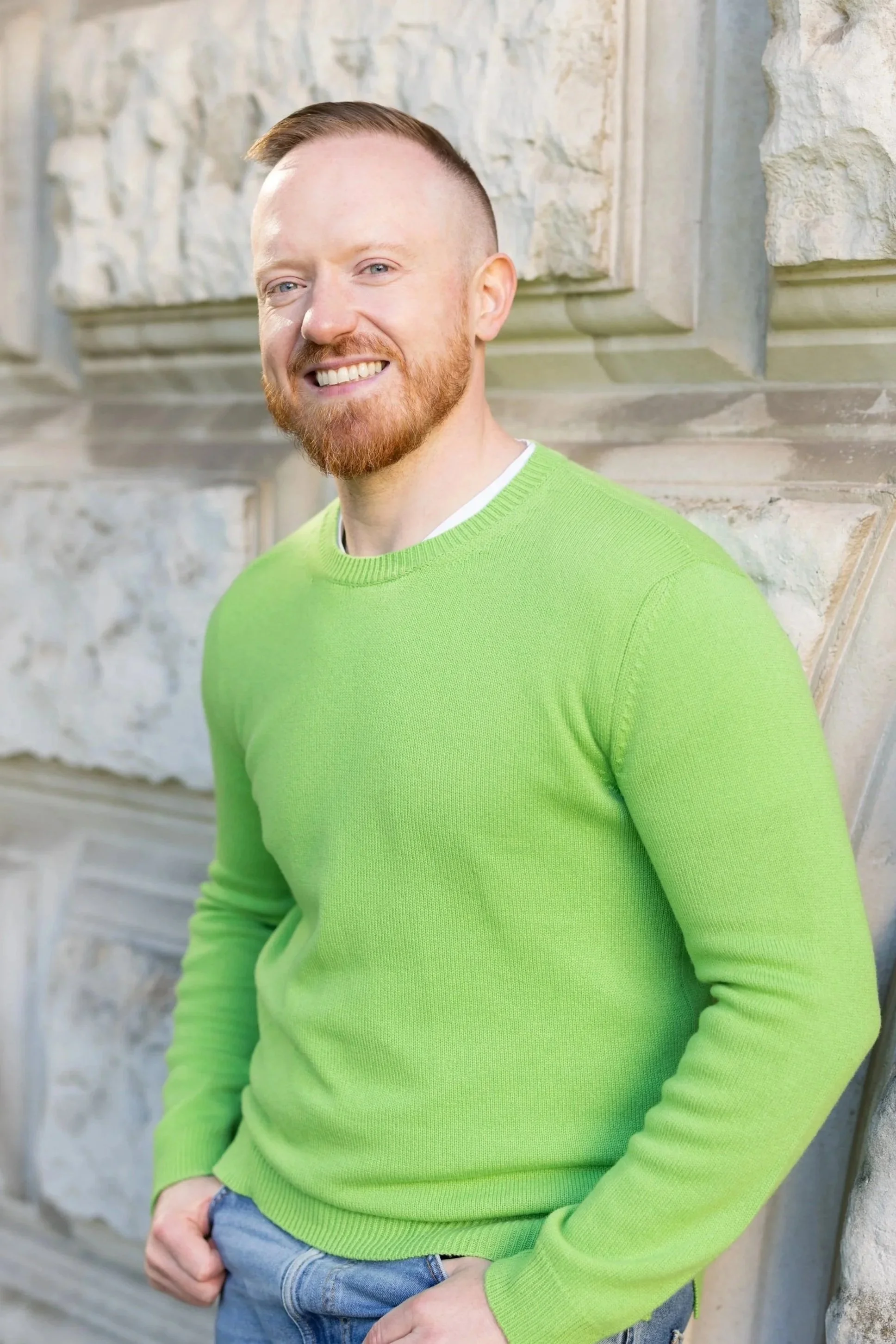 A young man with a red beard and short red hair wearing a bright green sweater and blue jeans, standing outdoors in front of a stone wall, smiling.