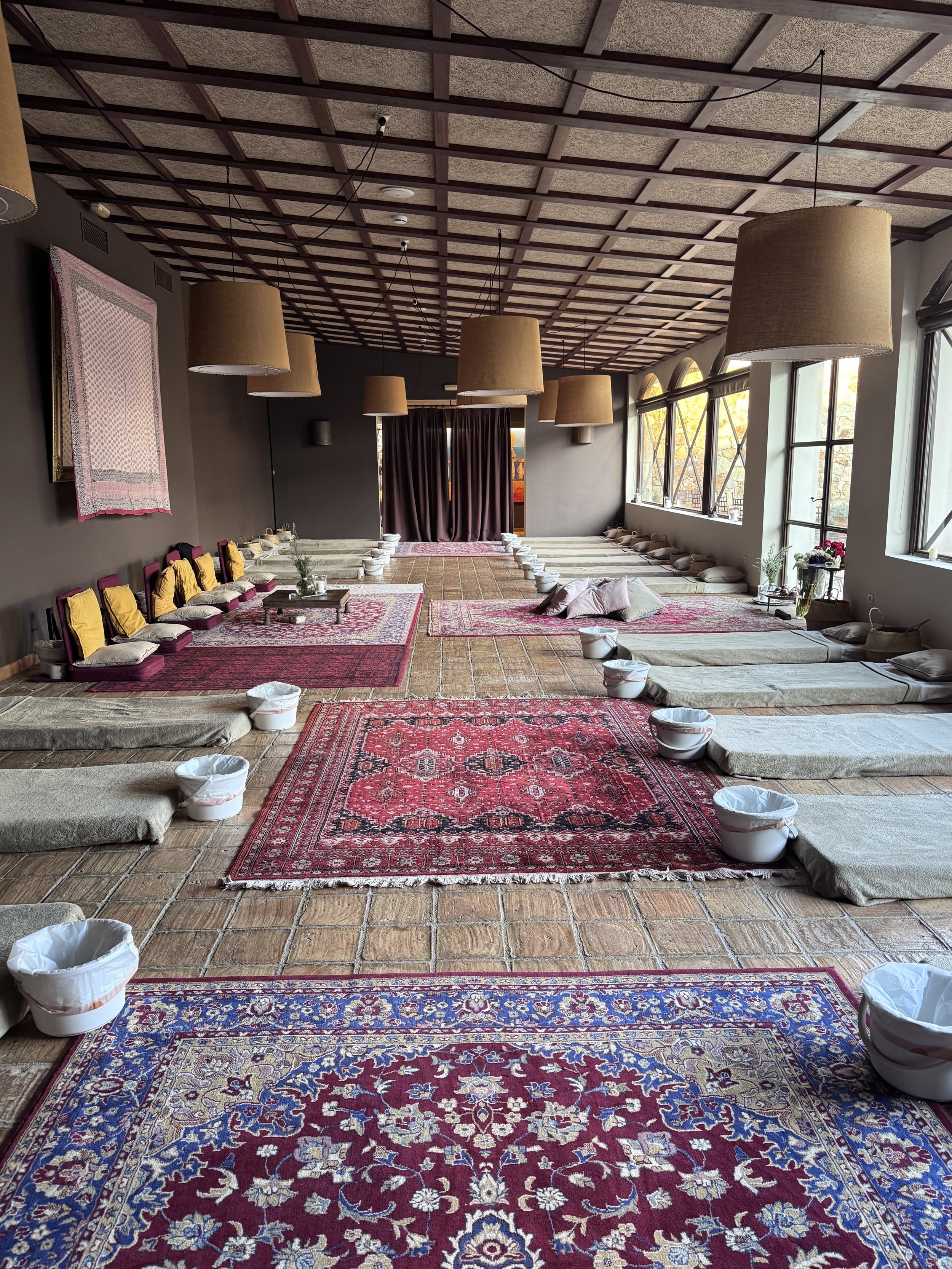 A large room with a high ceiling and windows along one side. The room is set up for a meditation or yoga session with cushions, rugs, and blankets arranged on the floor. There are hanging lamps and some potted plants.