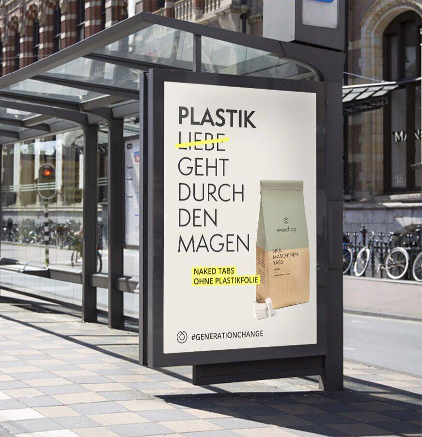 Plakat an einer Bushaltestelle, der mit buntem Text und einem beige Verpackungskarton beworben wird, das nachhaltige Produkte ohne Plastikfolie wie Nackte Tabs fördert.