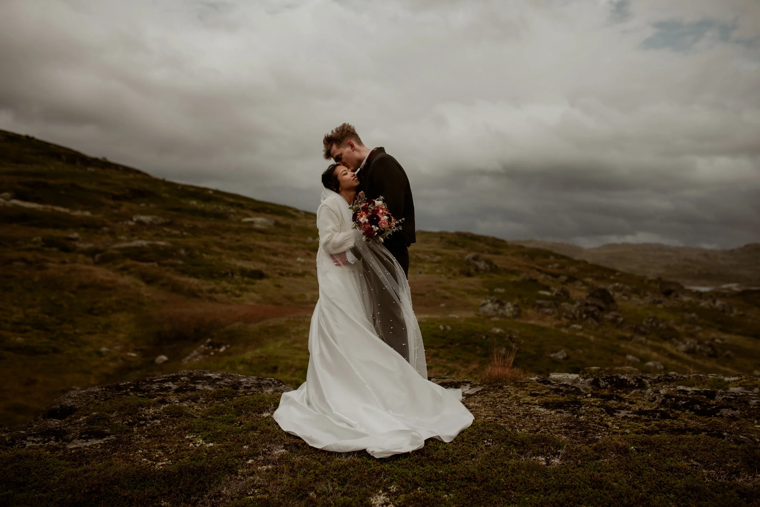 A bride and groom embrace outdoors on a cloudy day, standing on a mossy hilltop with rolling hills in the background. The bride wears a white wedding dress and holds a bouquet of flowers, while the groom wears a black suit.
