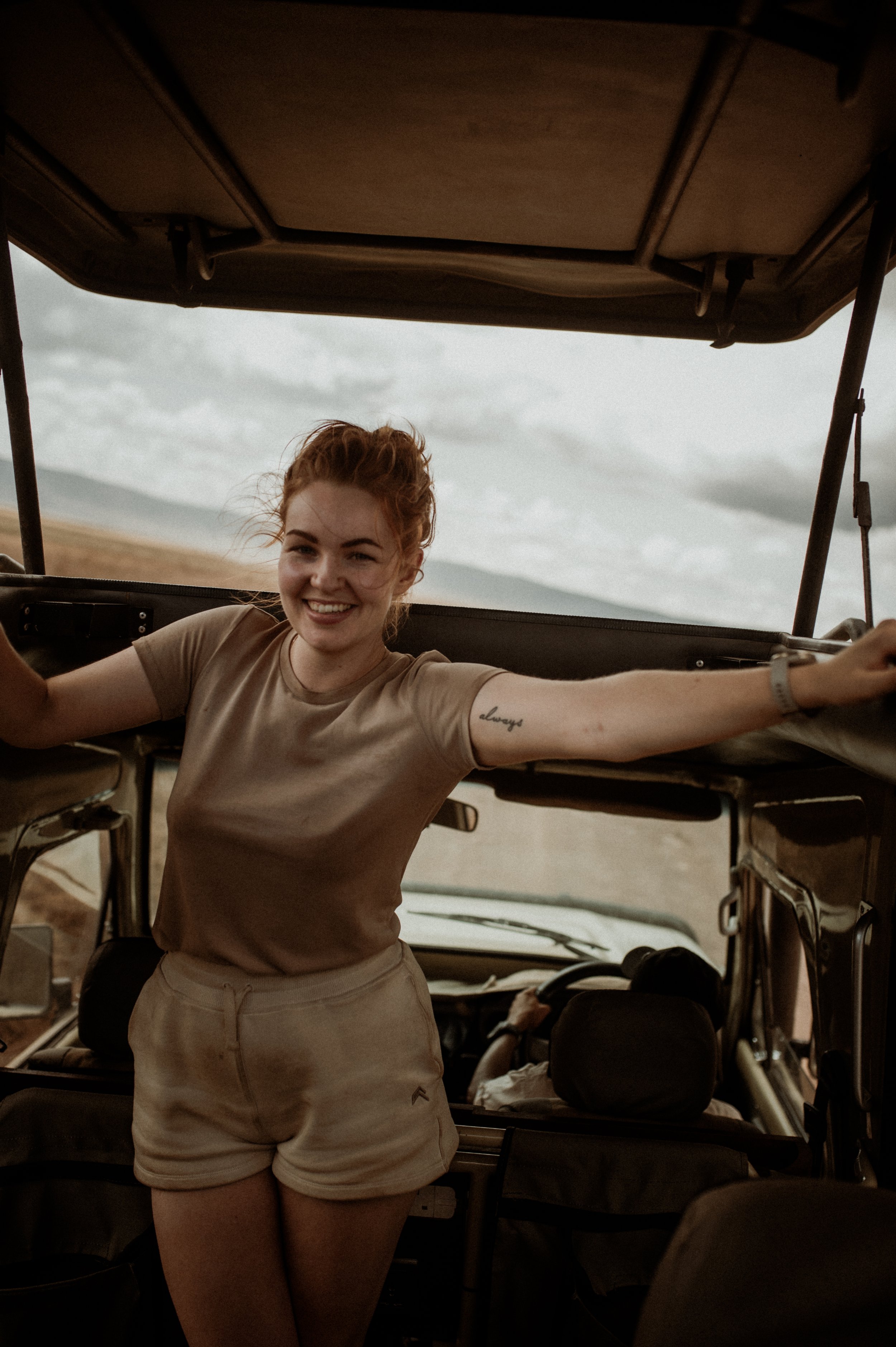 A smiling woman with curly red hair stands inside a vintage off-road vehicle with her arms outstretched, stretching across the width of the vehicle. The vehicle is in a rural, open landscape with cloudy skies.