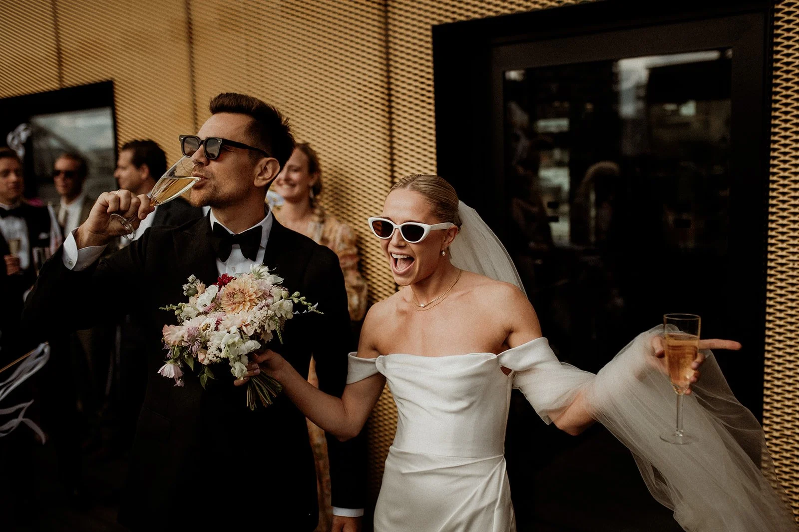 A bride and groom celebrating at their wedding reception, with the groom drinking champagne and the bride smiling, wearing sunglasses, holding a bouquet and a glass of champagne, surrounded by guests.
