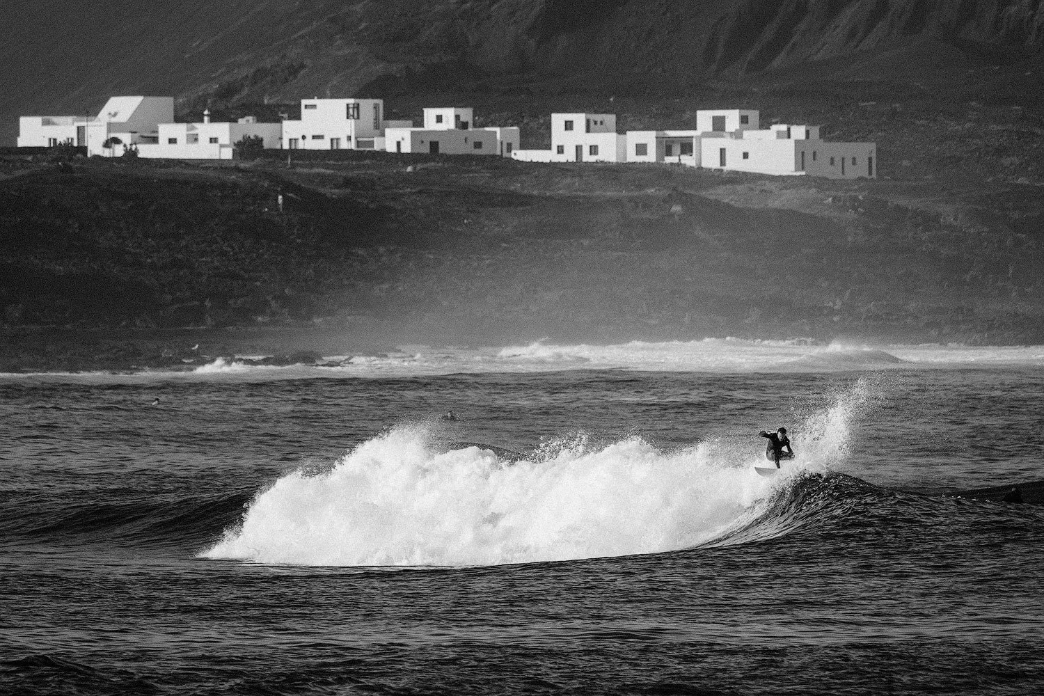 Black-and-white view of a surfer riding a breaking wave below a coastal village La Santa in Lanzarote and distant mountains.