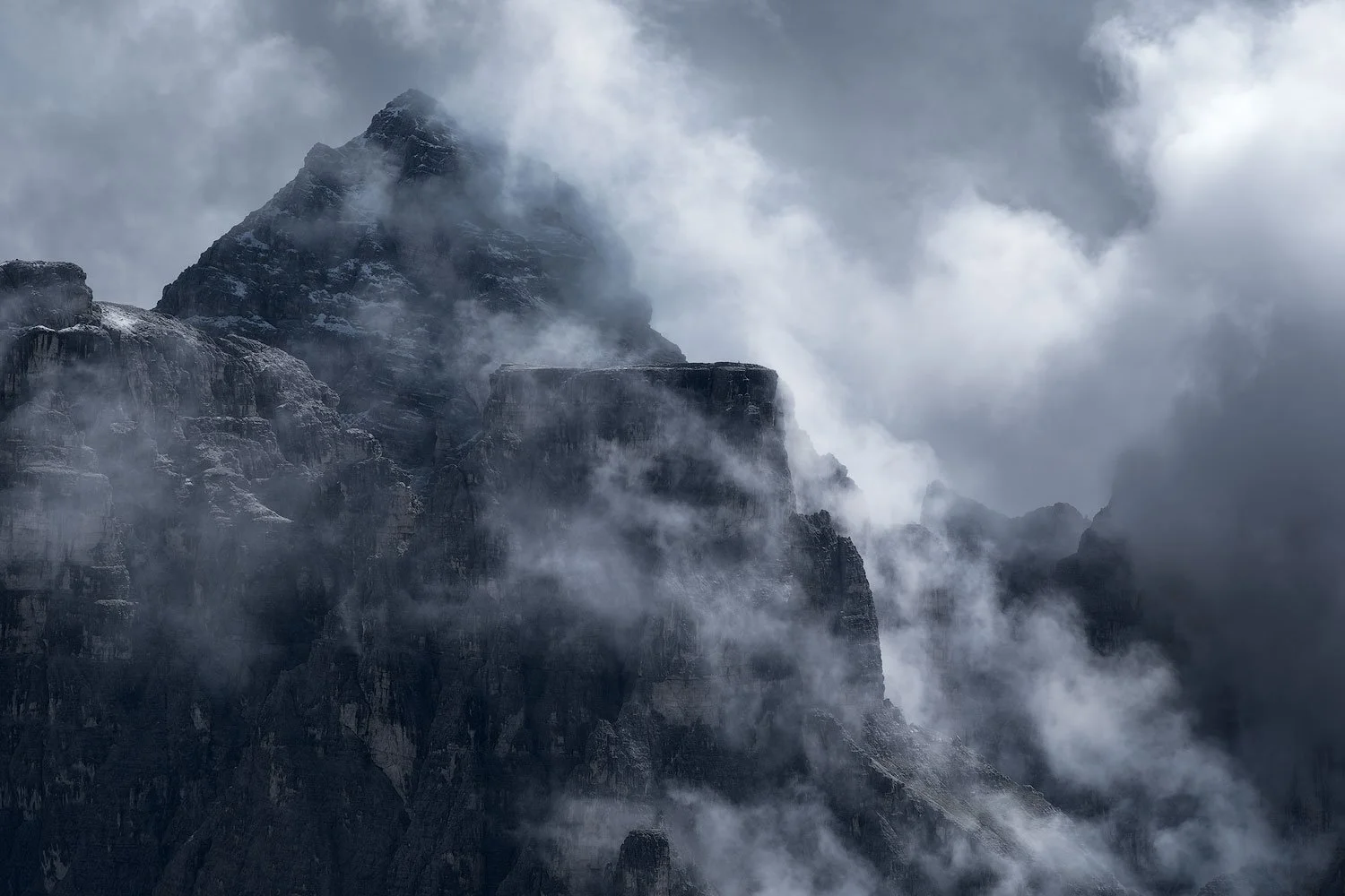 A tall mountain peak in the Italian Dolomites partially covered by fog and clouds, with dark rocky slopes.