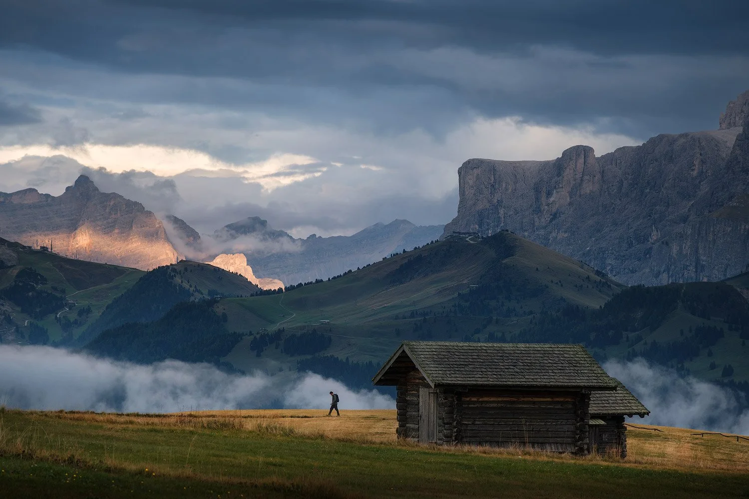 A wooden hut in the meadows of Alpe di Siusi beneath low clouds and mountain cliffs.