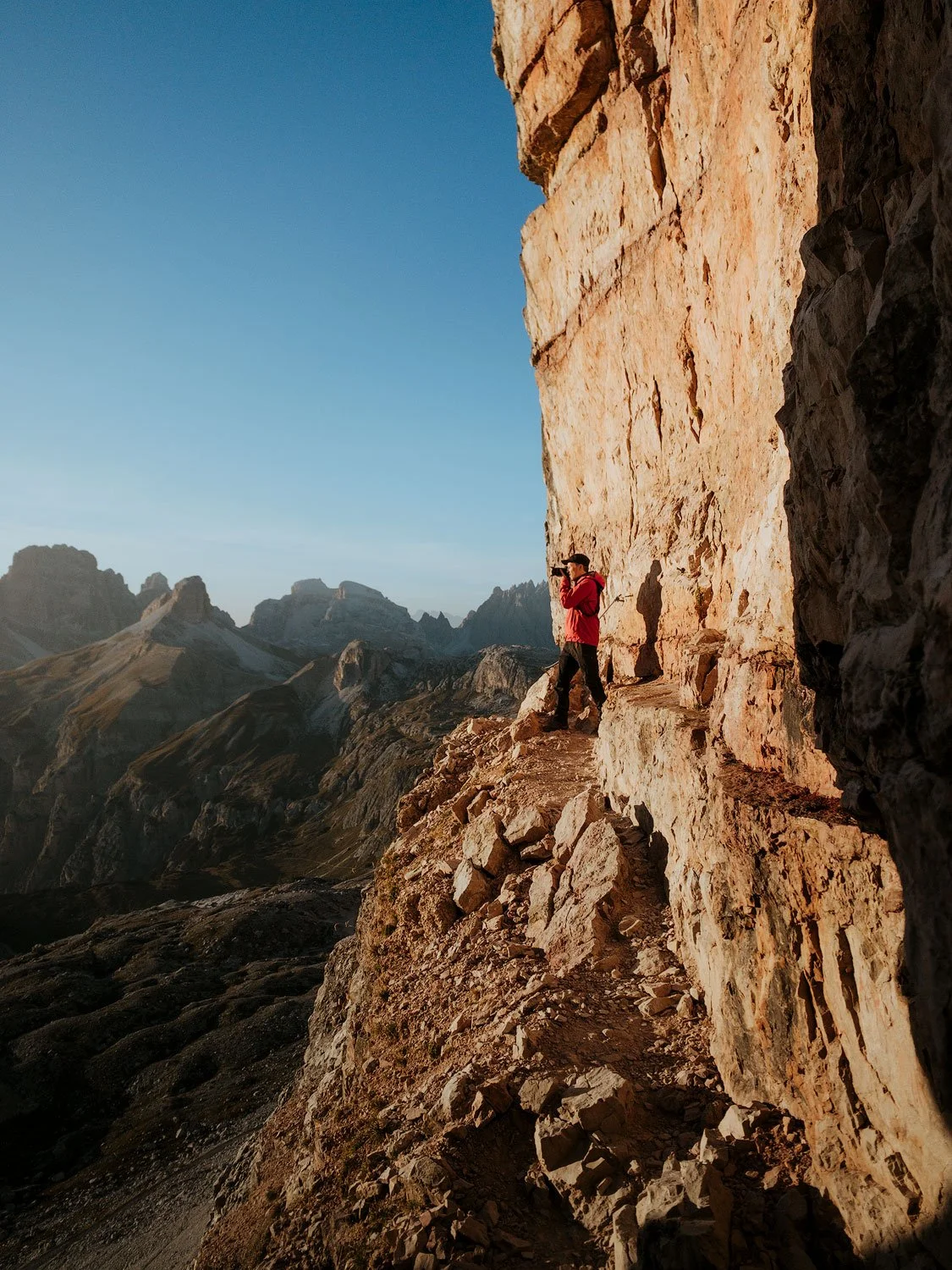 Person taking a photo on a mountain ridge in the Dolomites, with rocky cliffs and mountain peaks in the background during sunset.