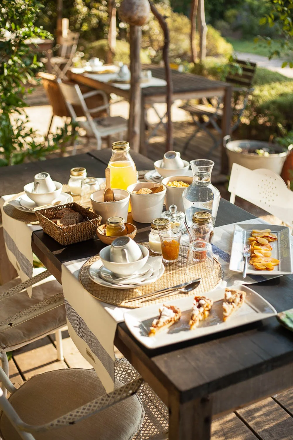 Outdoor breakfast table with plates of sliced fruit, pastries, bowls, jugs of juice and water, and coffee cups on a patio with trees and greenery.