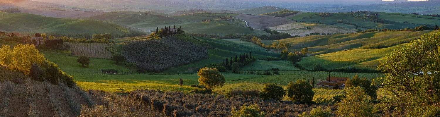 Warm directional sunrise light across the Tuscany countryside with glowing hills, olive groves, and layered shadows.