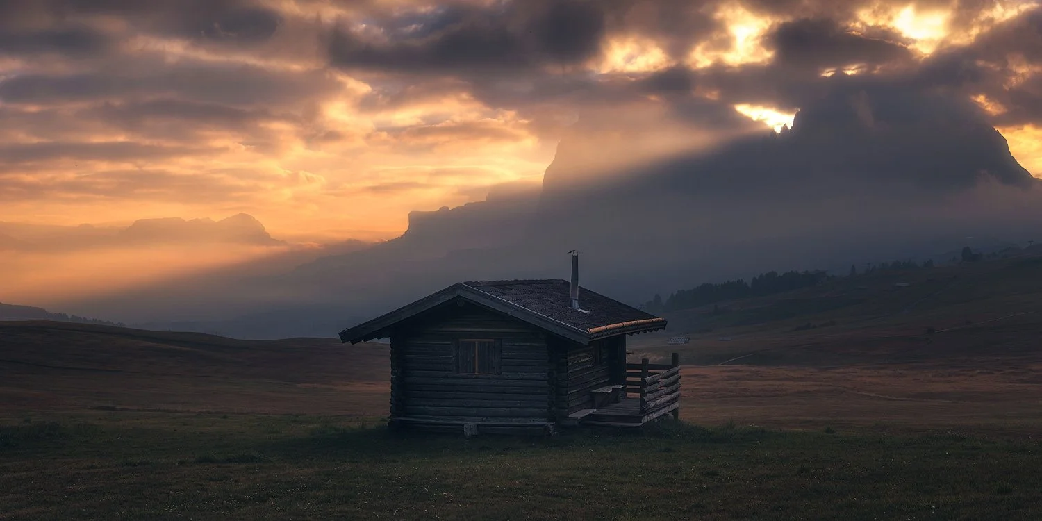 A wooden meadow hut silhouetted beneath orange dawn light and dark clouds at Alpe di Siusi.