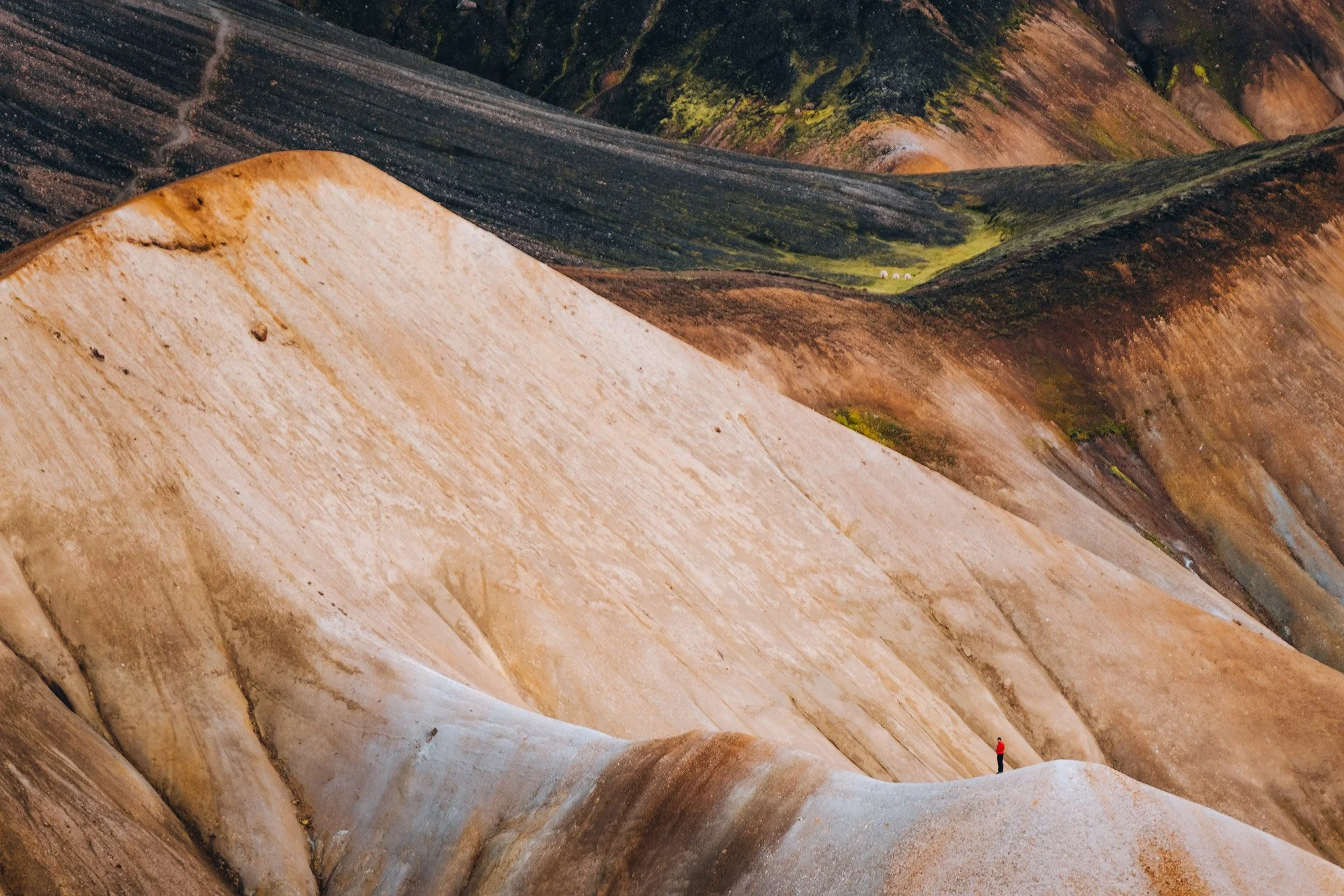 Lone person in red standing among layered ochre, white and green rhyolite slopes in the Icelandic Highlands, photographed with the Fujinon 50–140mm and 2× teleconverter.