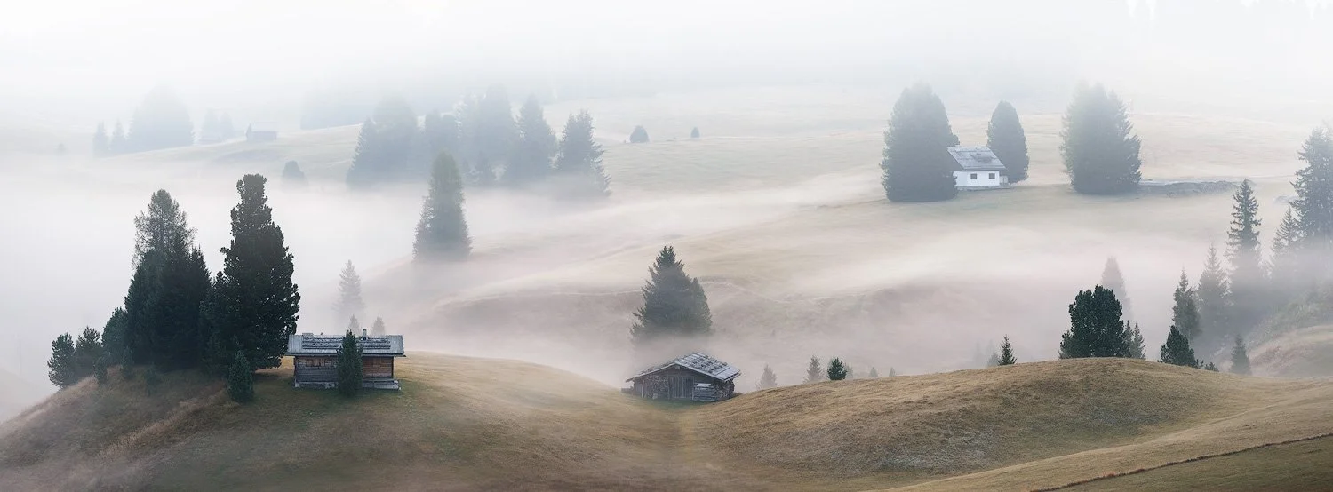 Panoramic xpan telephoto composition of wooden huts and conifers emerging through morning mist at Alpe di Siusi.