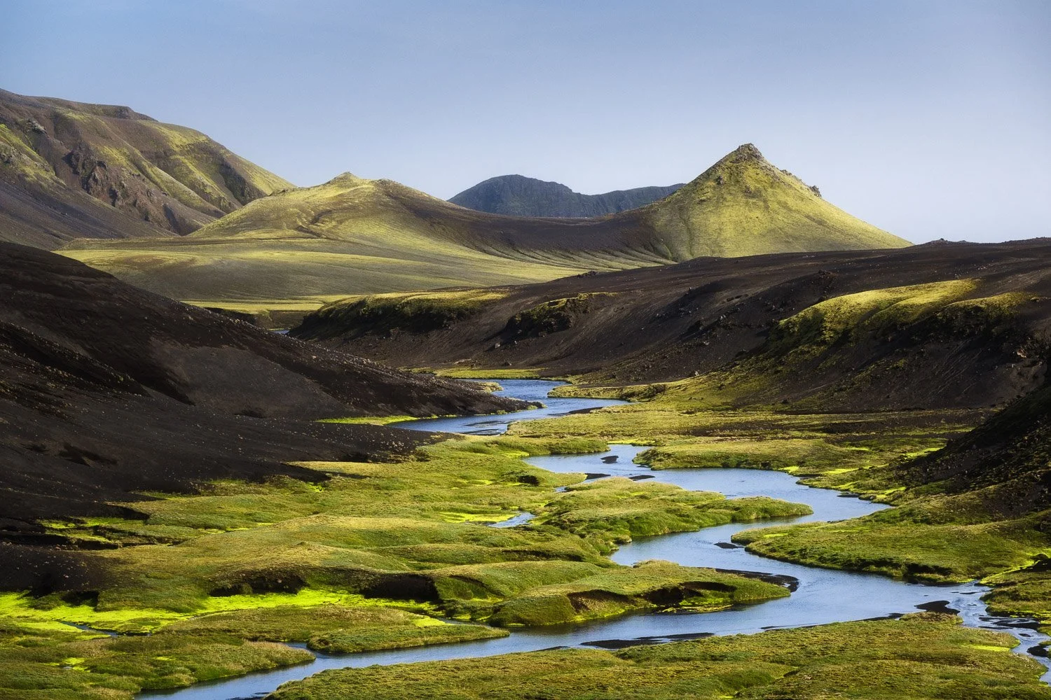 Winding river flowing between green moss and black volcanic hills beneath sunlit mountain peaks in Mælifellssandur, Iceland.