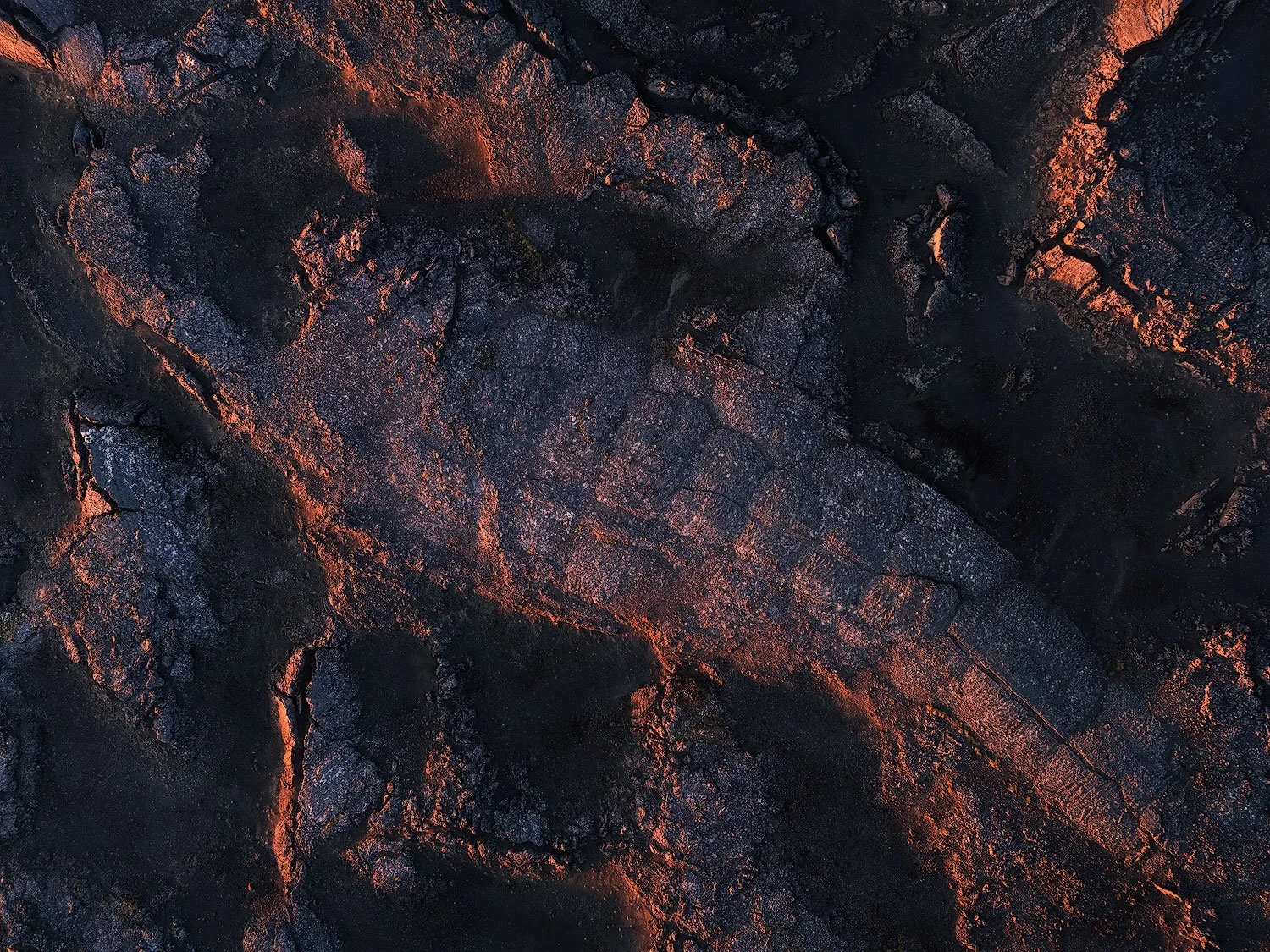 Close-up of cooled volcanic lava with dark, cracked, and textured surface illuminated by subtle reddish glow.