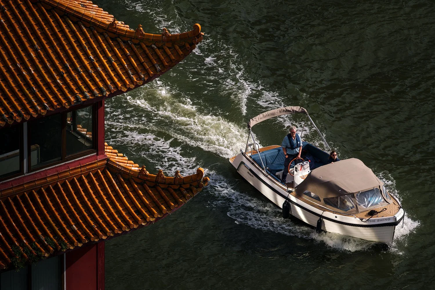 Small motorboat passing a traditional red-and-gold roof on a Rotterdam waterway, viewed from above.