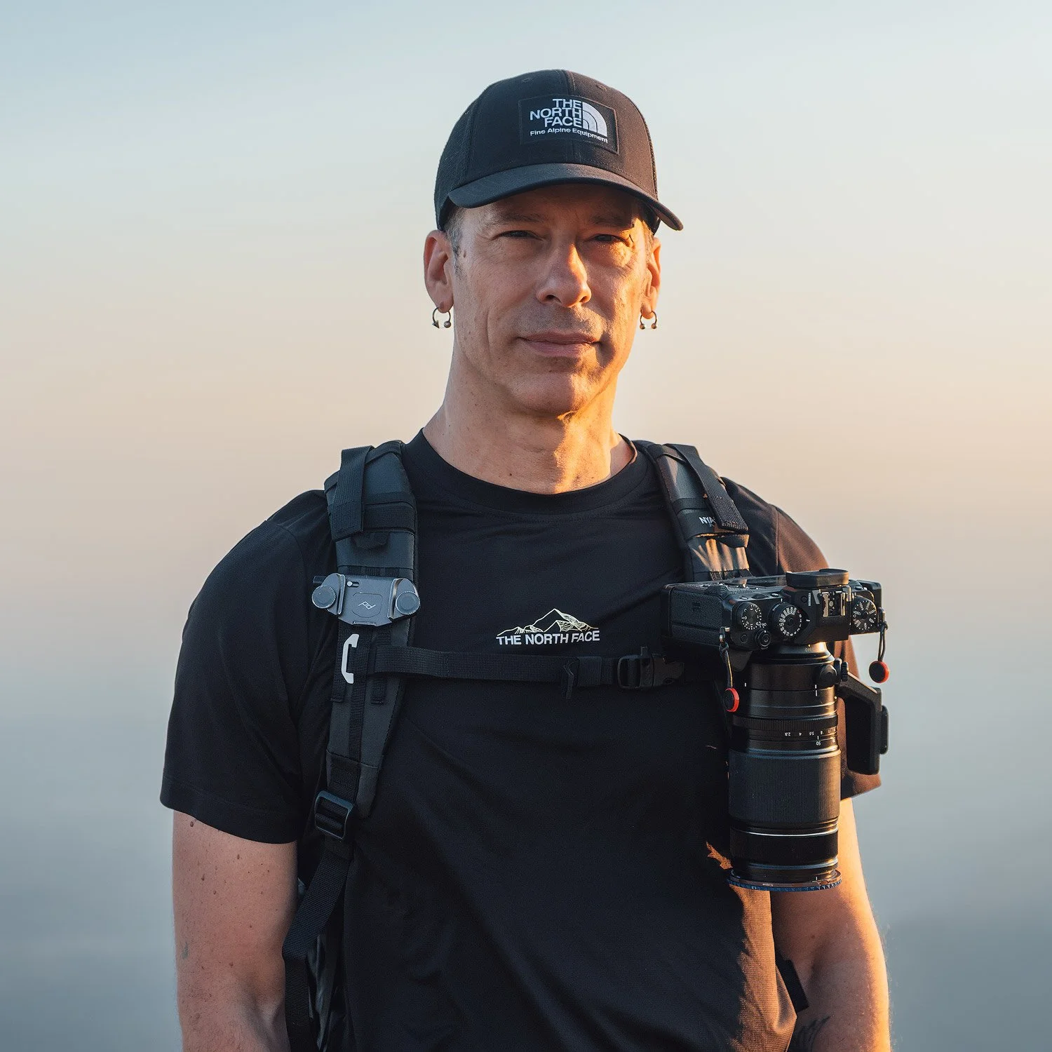 andrea livieri portrait with the nya-evo backpack, the north face hat and t-shirt, and a fujifilm camera