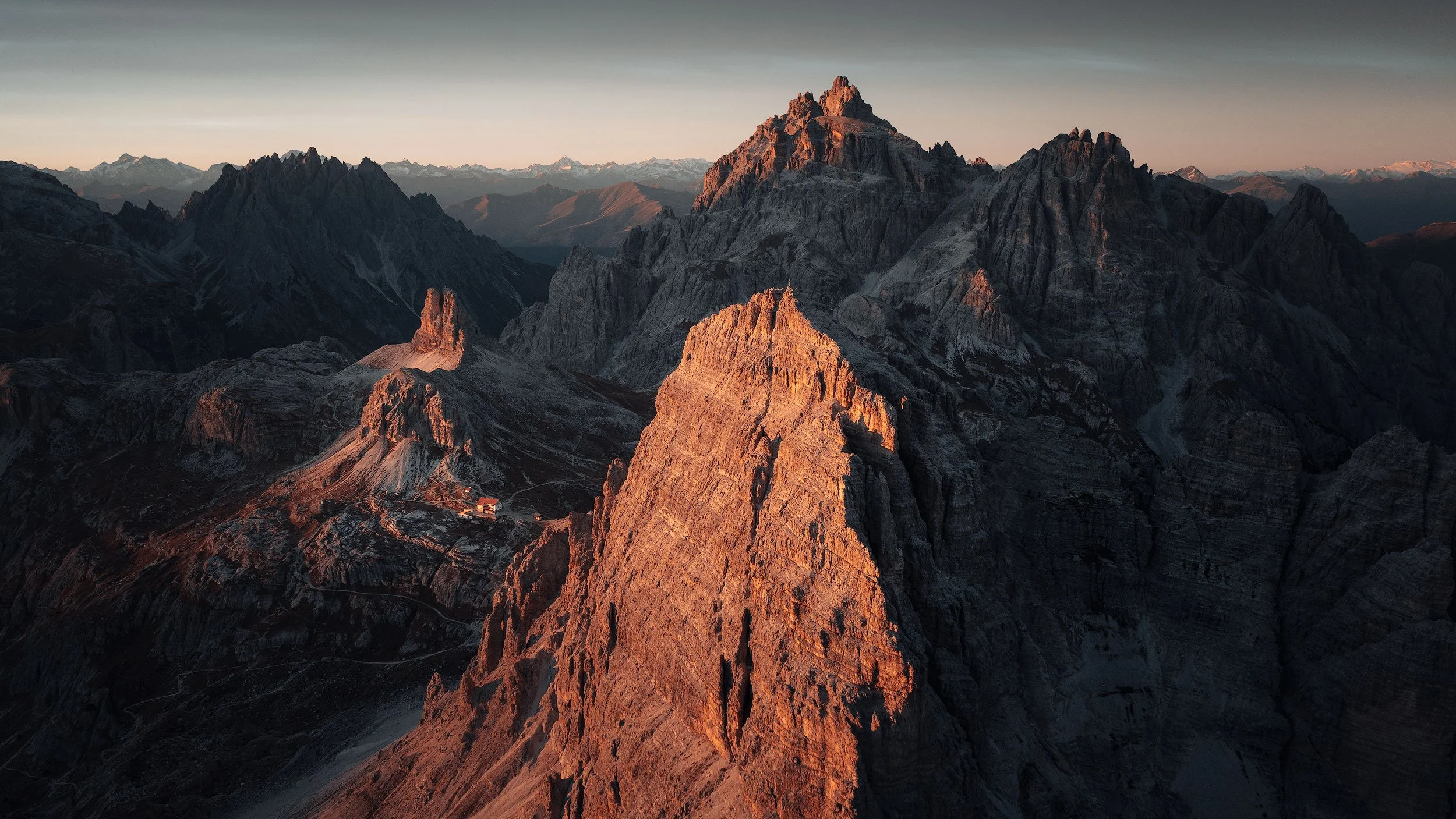 Sunset over rugged tre cime di lavaredo mountain peaks in the dolomites with rocky terrain and a few small structures at the base.