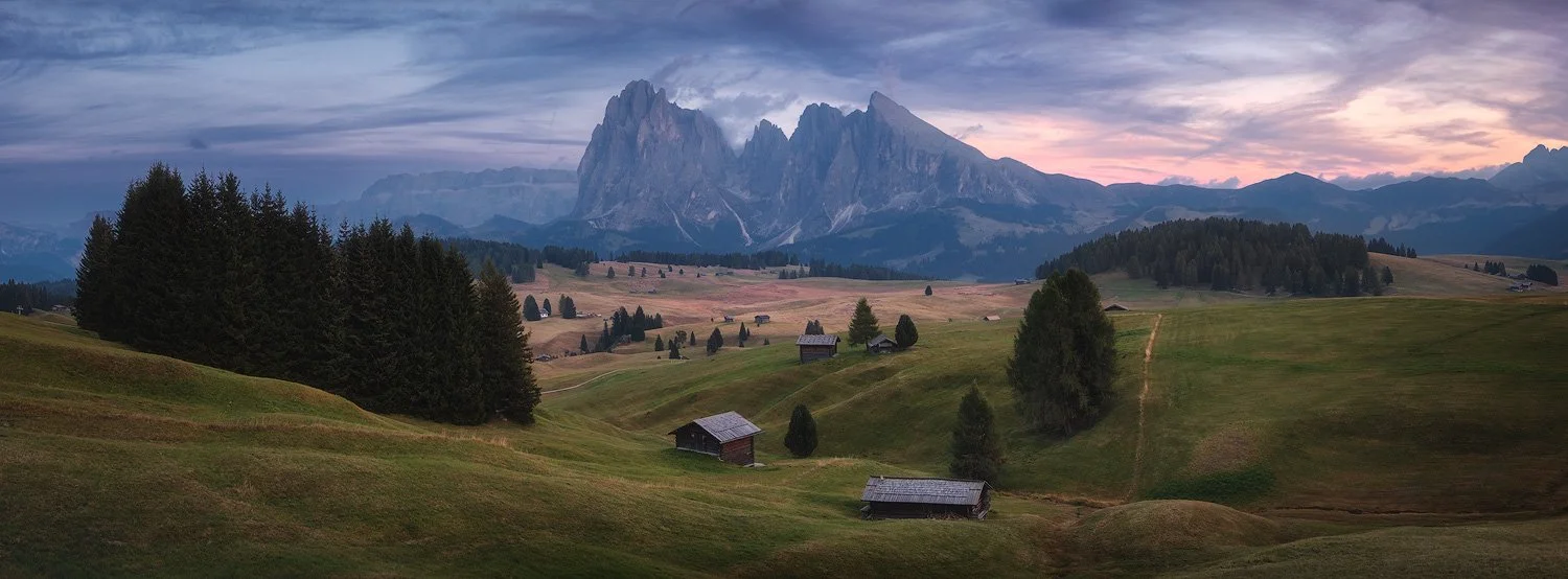Wide panoramic xpan photograph of Alpe di Siusi’s meadows, wooden huts, and Sassolungo beneath a pink evening sky.