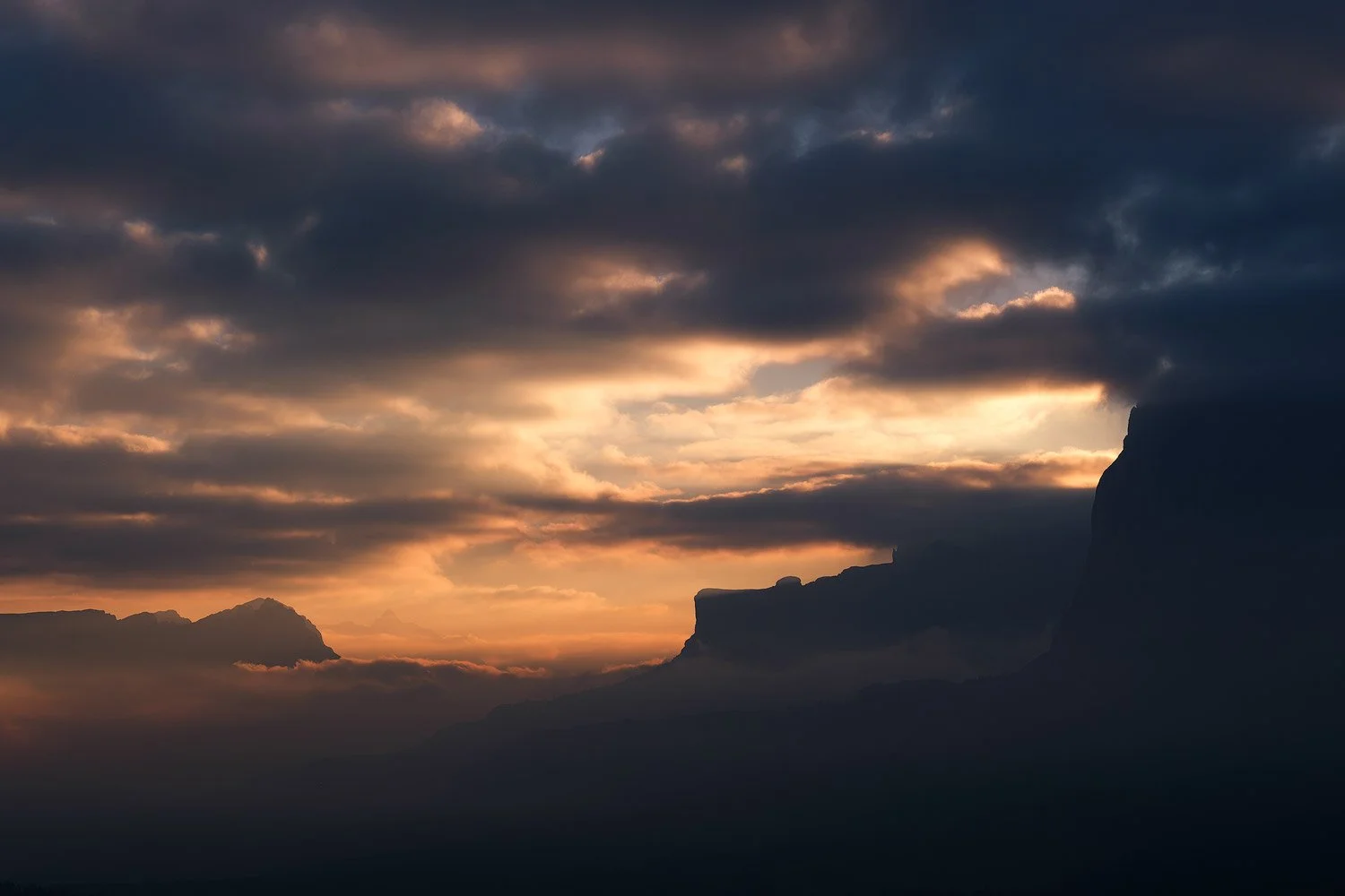 Warm dawn light breaking through heavy clouds above the mountain silhouettes surrounding Alpe di Siusi.