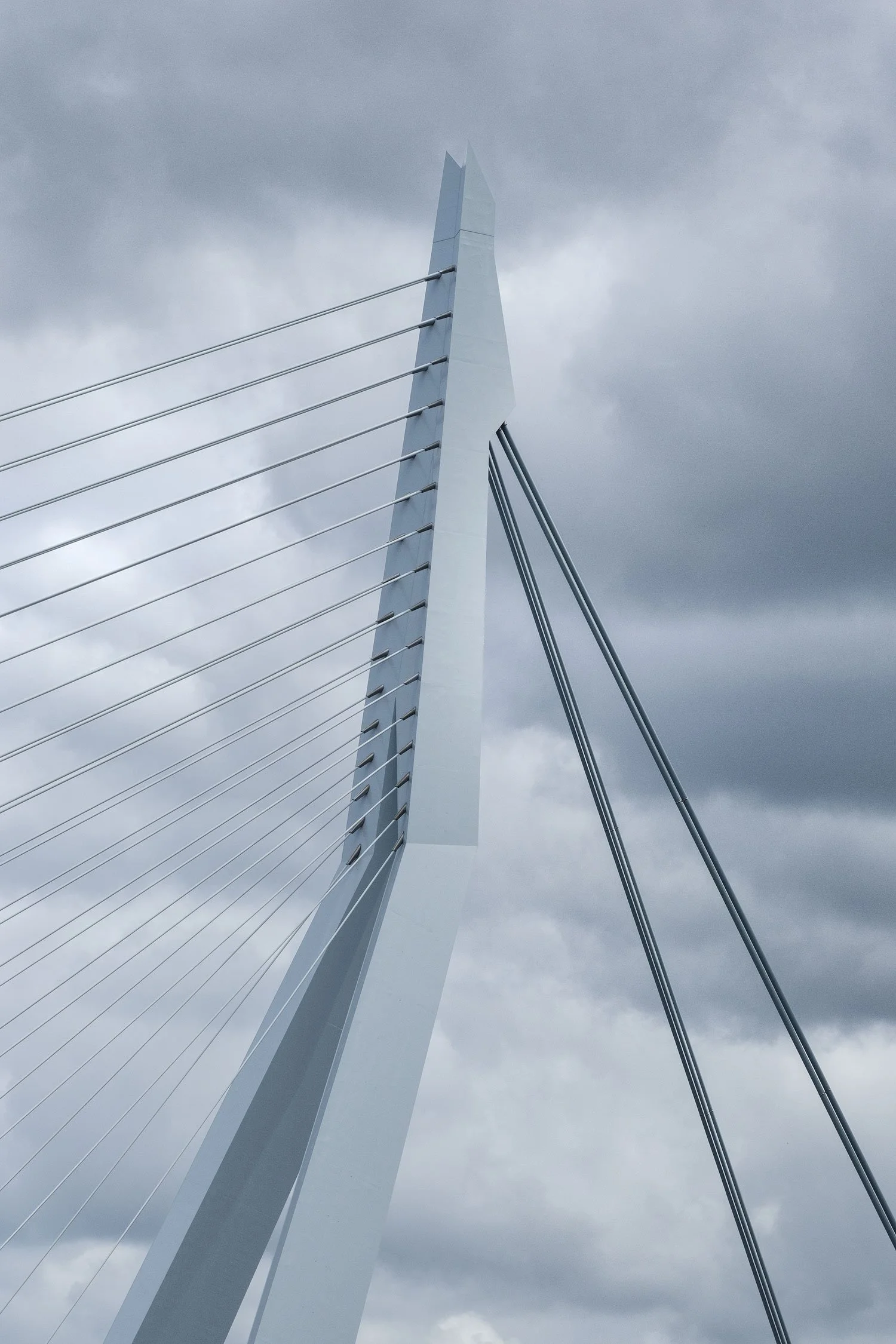 Upward telephoto view of the Erasmus Bridge pylon and cables against stormy clouds in Rotterdam.