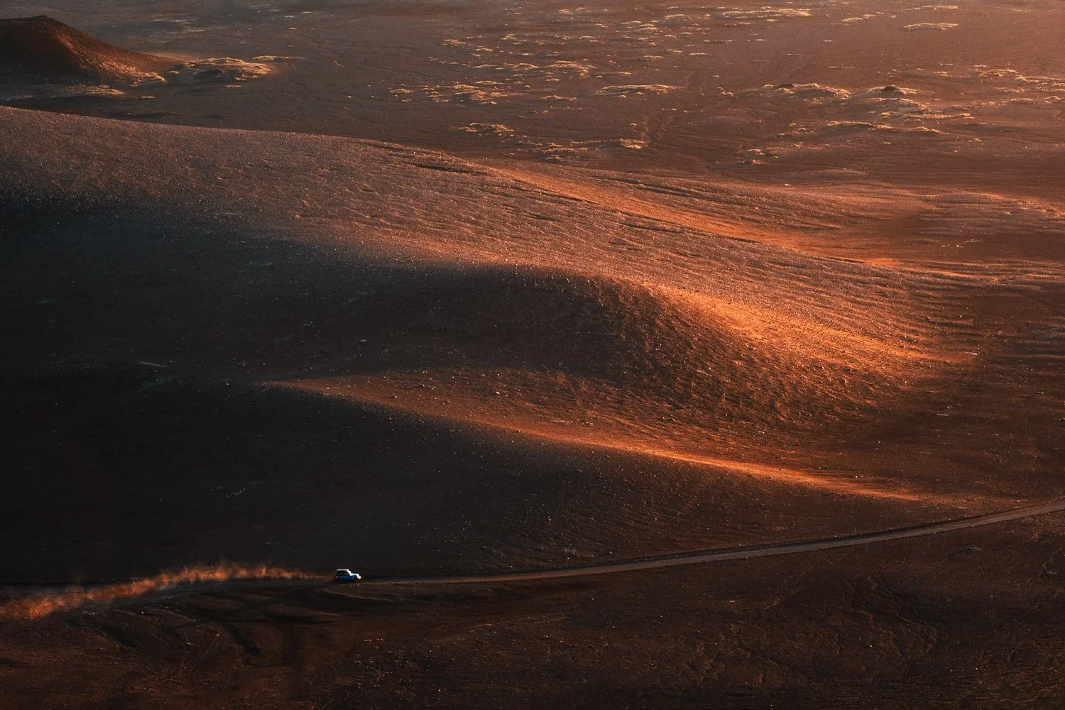 Small vehicle driving along a road through a vast dark volcanic landscape illuminated by warm low-angle light.