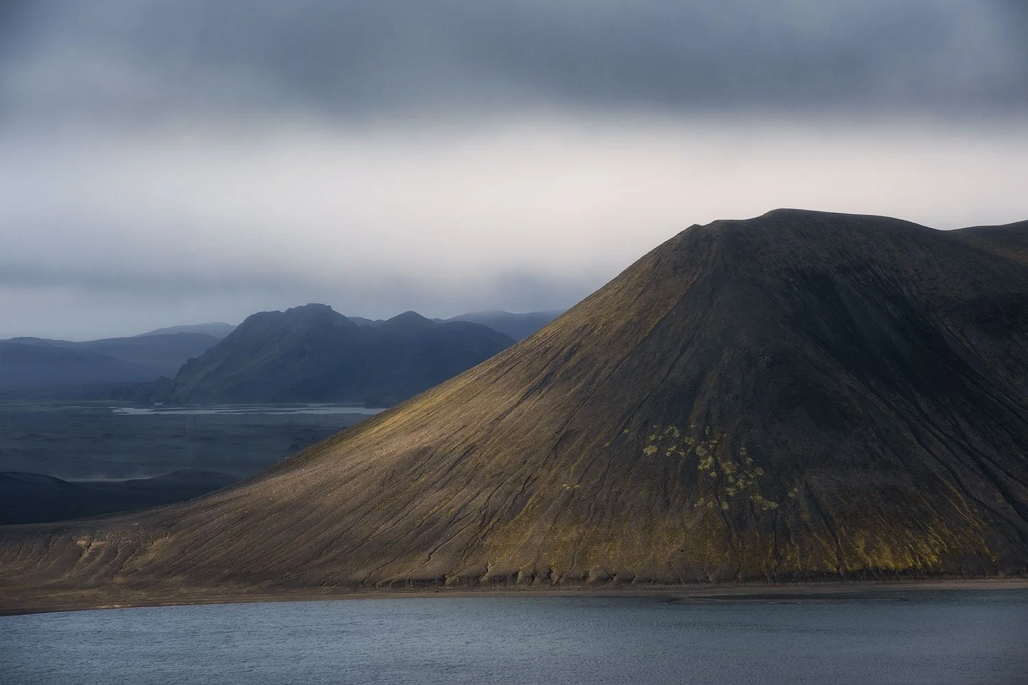 Sunlit green volcanic hills, stream channels and a small lake beneath a dark ridge in the Icelandic Highlands.