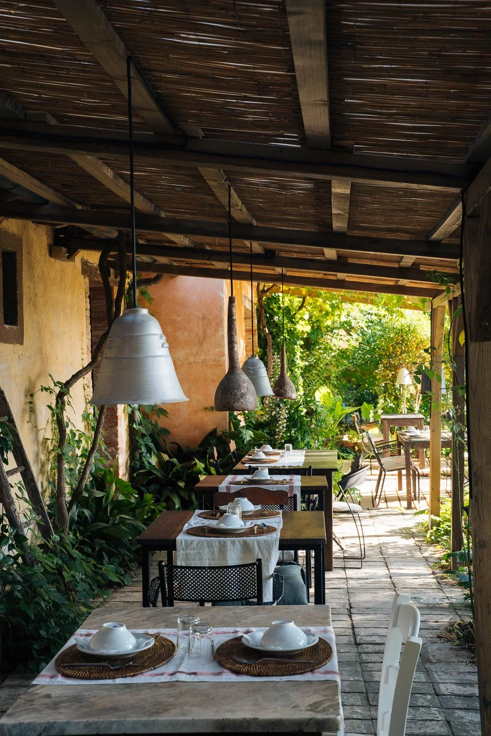 Outdoor restaurant seating area with tables, chairs, and hanging pendant lights under a wooden roof, surrounded by greenery and sunlight.