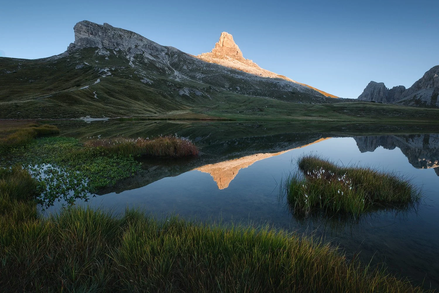 A sunlit peak reflected between grassy islands in the still water of Laghi dei Piani.