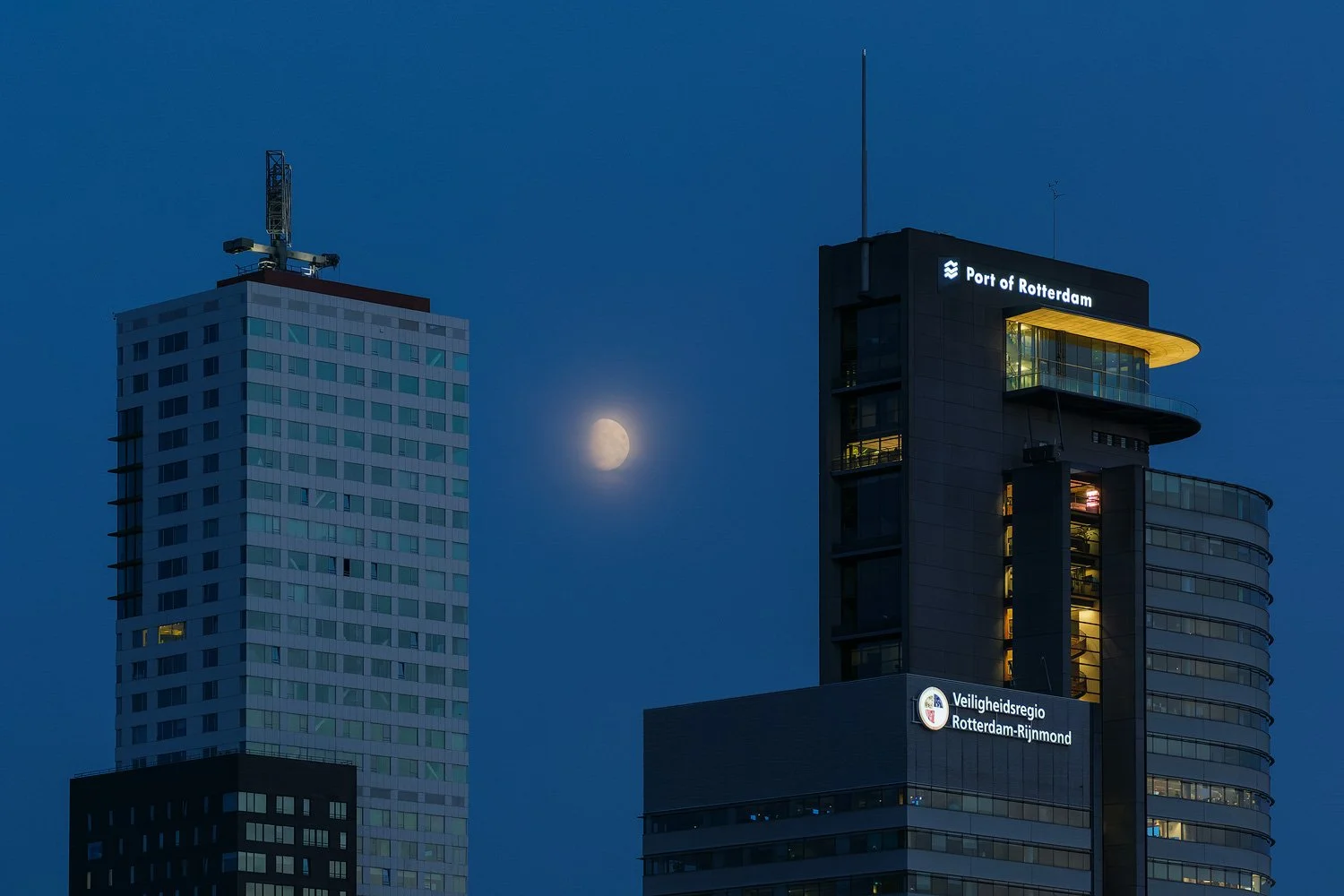 Moon rising beside illuminated high-rises and the Port of Rotterdam headquarters during blue hour.