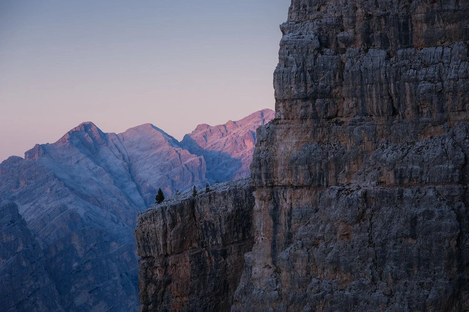 A shaded rock tower at Cinque Torri overlooking distant Dolomite peaks in pink evening light.