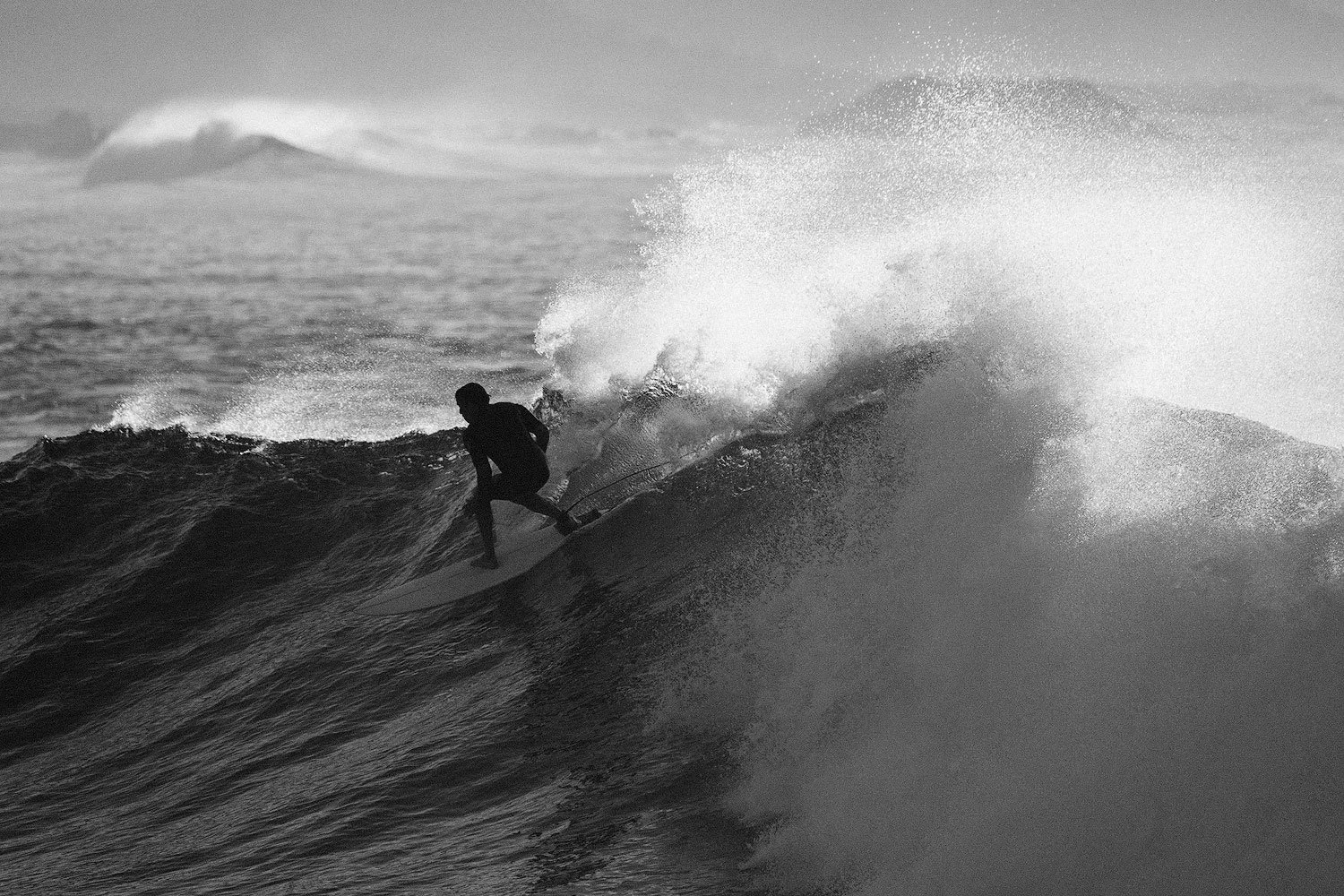 Black-and-white photograph of a surfer riding a steep wave as backlit spray breaks above him.