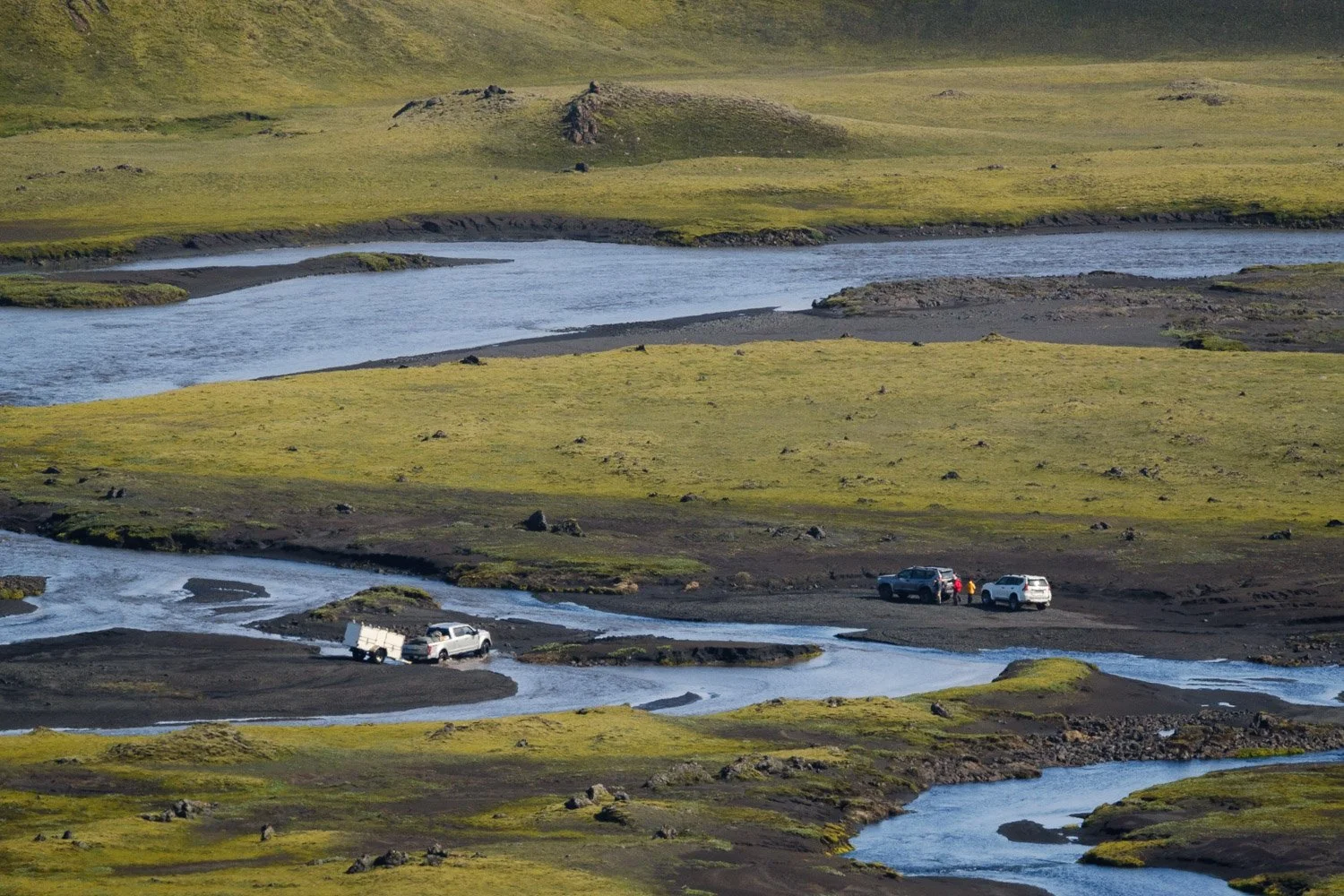 Three 4x4 vehicles beside the Hólmsá braided river crossing through moss-covered volcanic terrain in Mælifellssandur, Iceland.