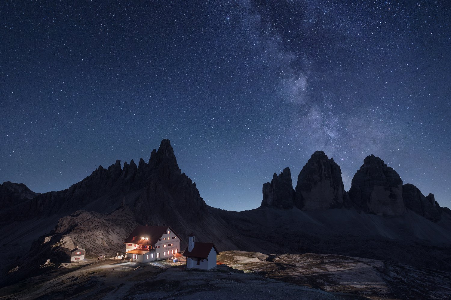 The Milky Way above the silhouetted peaks of Tre Cime di Lavaredo and the illuminated mountain hut.