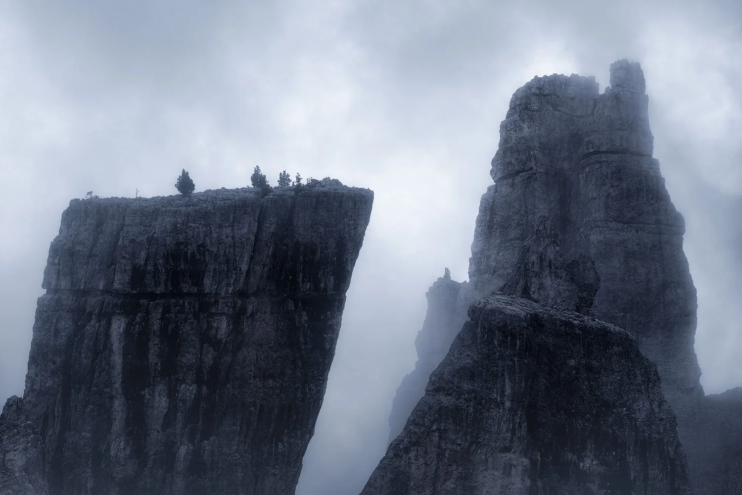Tall, rugged rock formations with sparse trees on top, shrouded in fog and clouds in the Dolomites