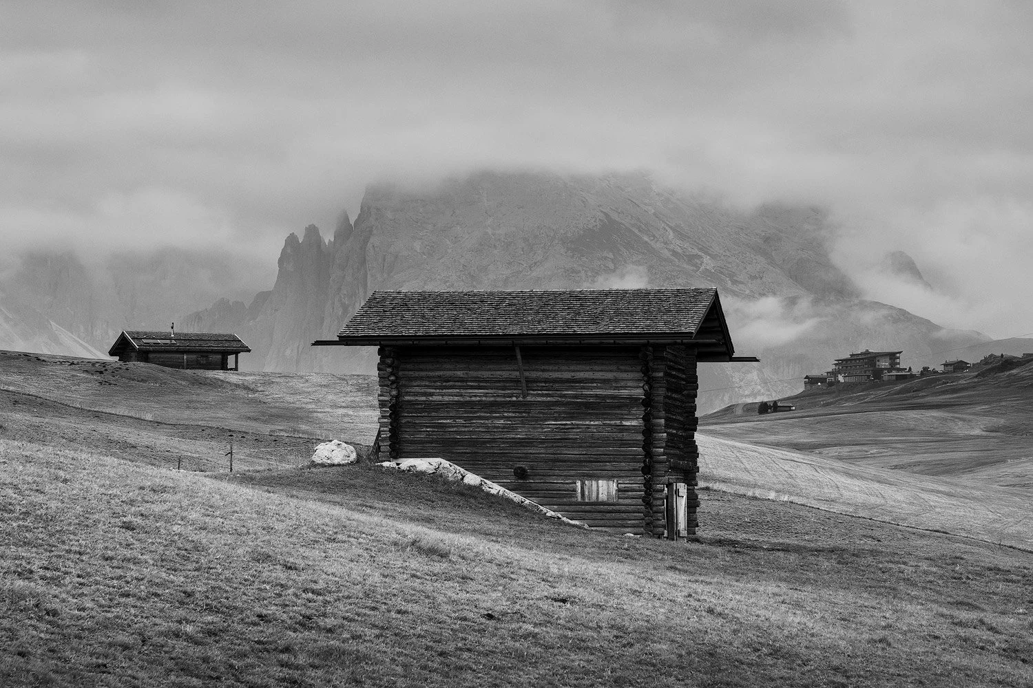 Black-and-white composition of wooden huts on rolling meadows beneath cloud-covered mountains at Alpe di Siusi.