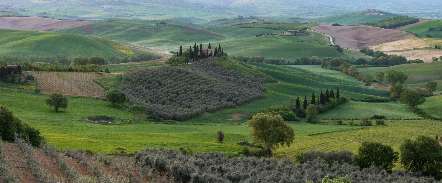 Wide panoramic photograph of Val d’Orcia in Tuscany captured before sunrise with soft atmospheric light and layered green hills.