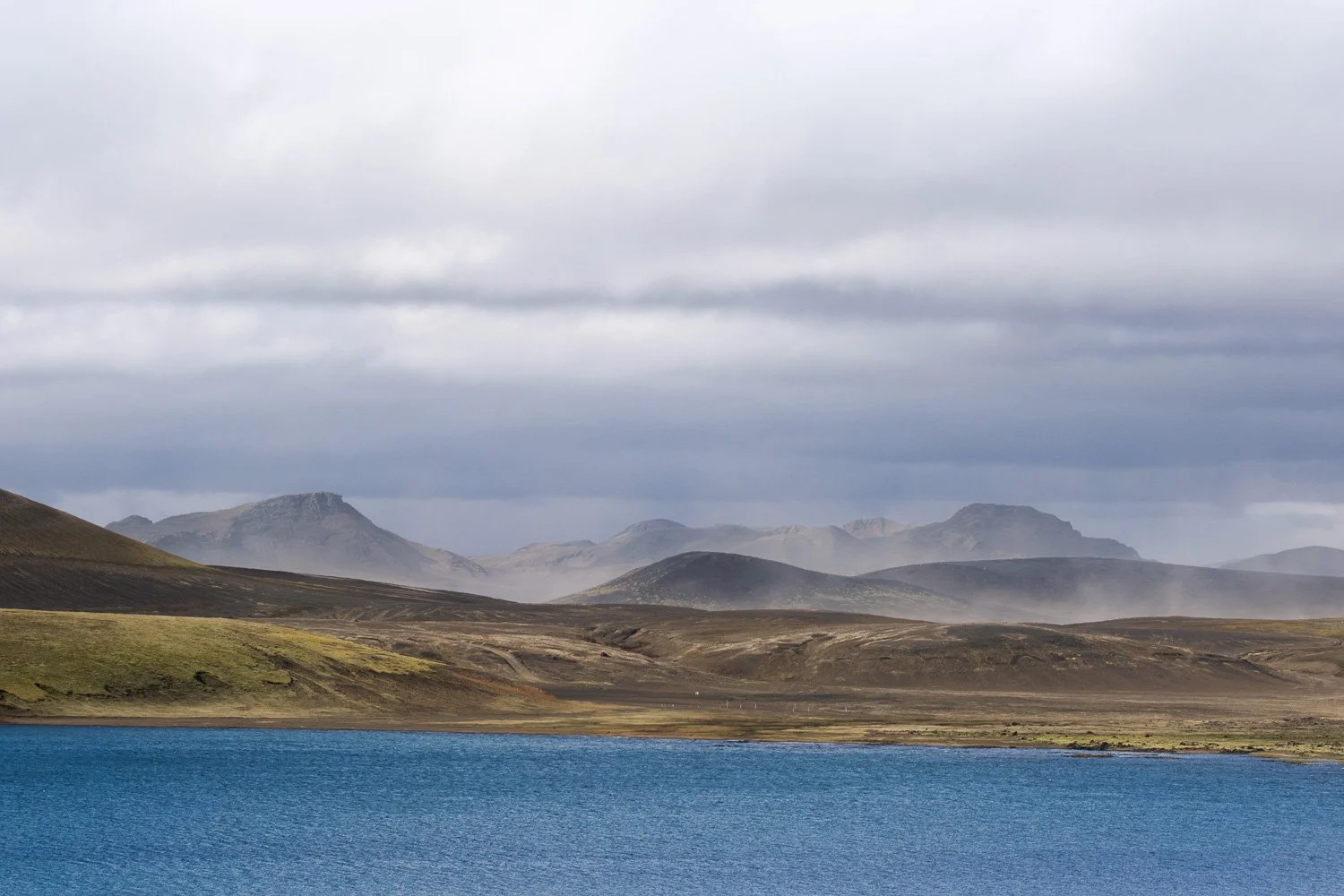 Wide view of a blue lake, volcanic mountains, blowing sand and cloudy sky in the Icelandic Highlands.
